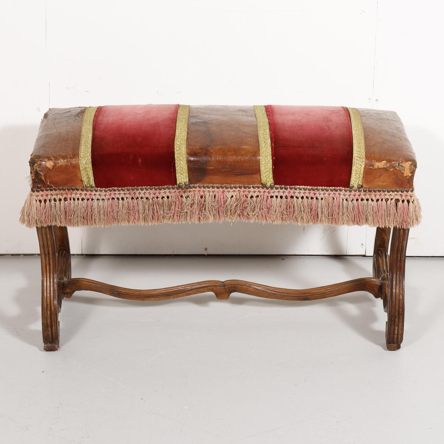 19th century Spanish Louis XIV style bench or stool having original leather and red velvet upholstery with metallic trim tape and tri-colored brush fringe trim, circa 1890s. This handsome backless bench is raised on hand carved walnut os de mouton