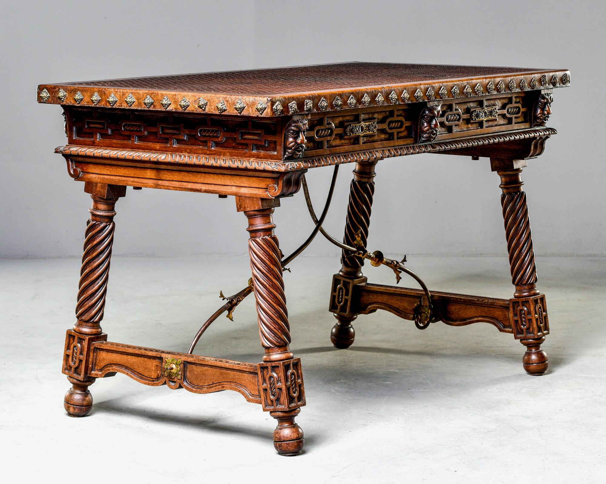 Spanish library or writing table features a new, embossed leather top, decorative brass nail head studs on the desktop edges, carved faces on the apron and two functional locking drawers, circa 1870s. Legs and supports all have carved details and
