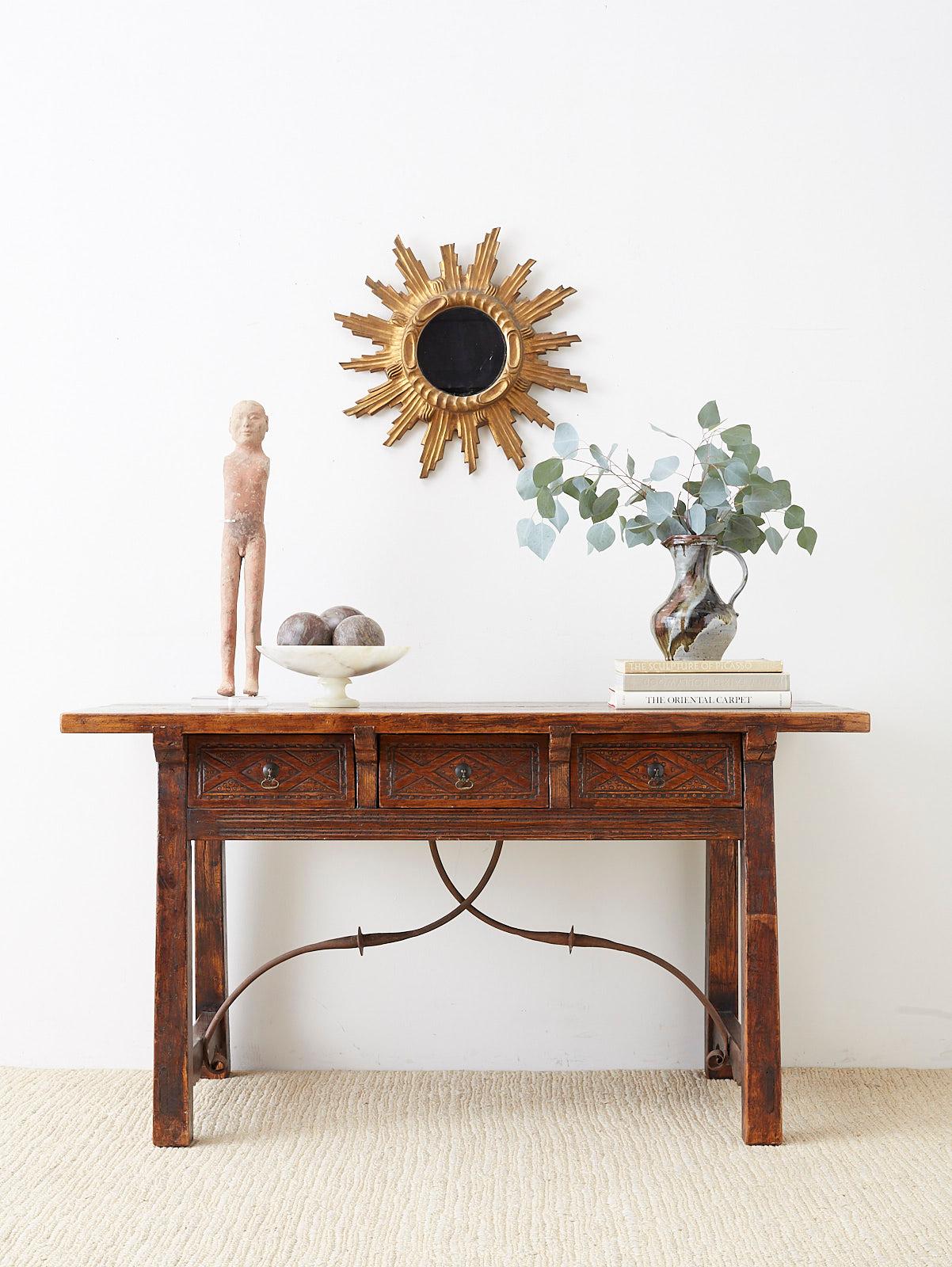 Rustic 19th century Spanish library table or writing table featuring iron trestle supports. Made in the Baroque taste fronted by three storage drawers with decorative carved fronts and iron pulls. Constructed with mortise and tenon joinery having a