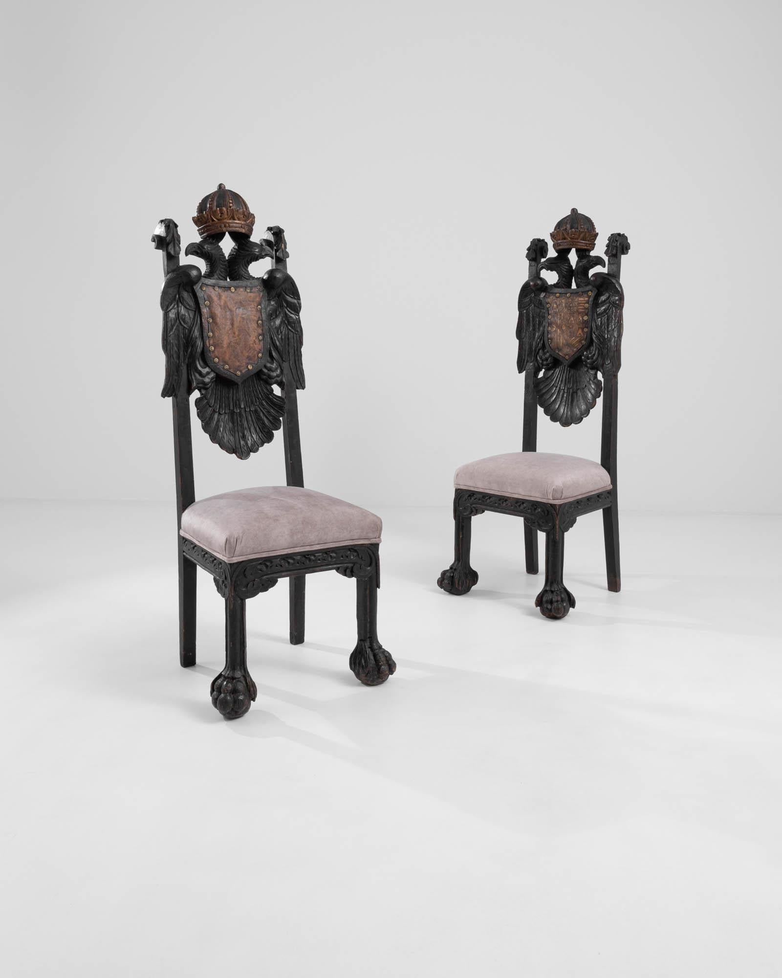 A wooden pair of upholstered armchairs crafted in 19th century Spain. Twin eagles, perched across the back of these exquisite chairs, share a coat of arms, capped in a regal helm. The dark hardwood that composes these chairs' structure has been