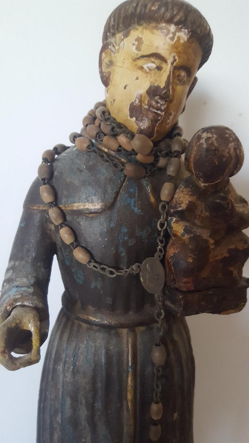 This Mexican Colonial figure of St. Anthony holding the Christ Child is made of carved wood with a blue/black, cream and gold painted finish that is highly distressed and worn from age and use. It is adorned in a wood and metal Rosary.