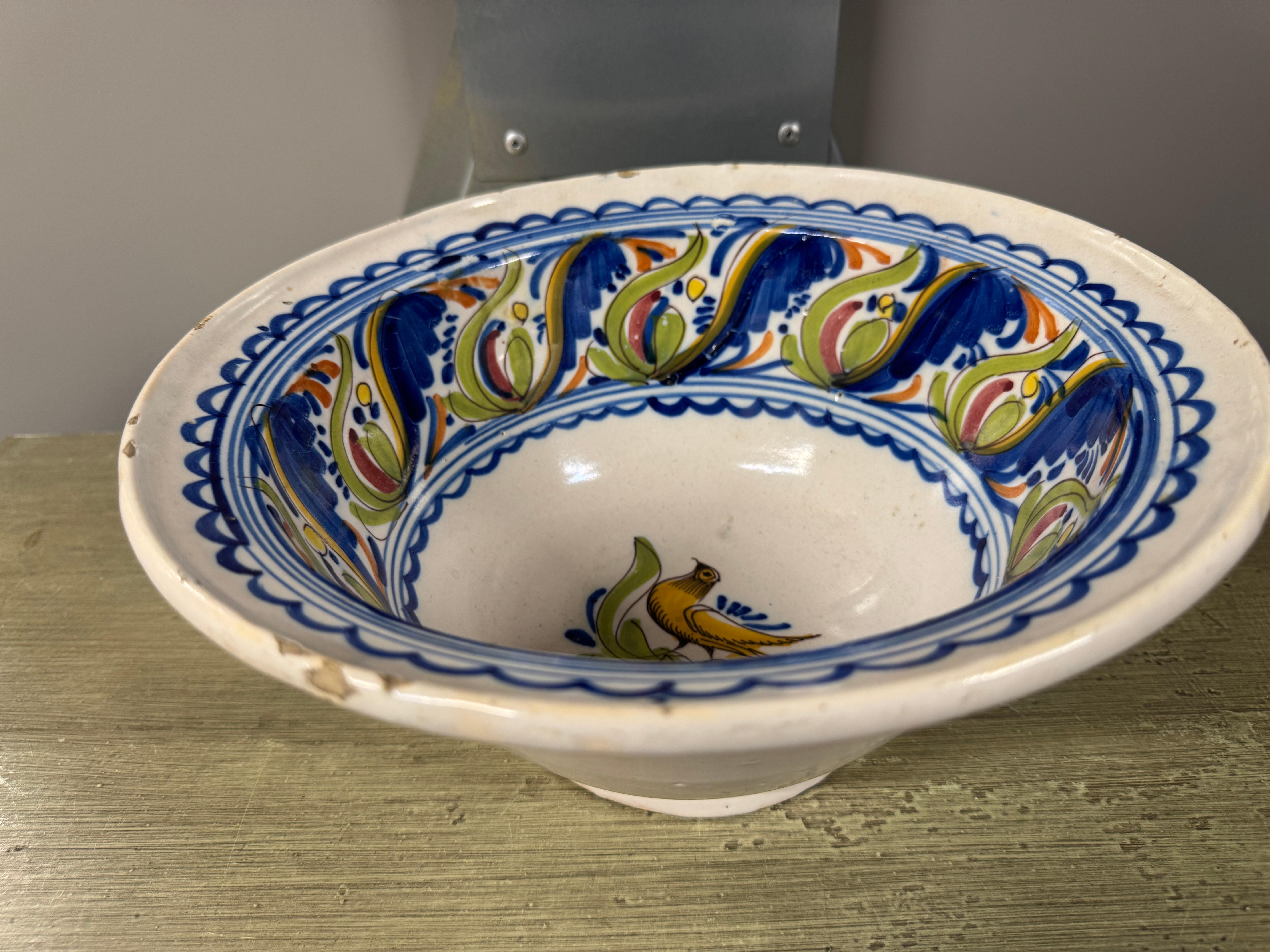 Cuenco español Manises del siglo XIX, presenta una base blanca con motivos azules, verdes, amarillos y naranjas, incluido un pájaro estilizado y elementos florales alrededor del borde. 

