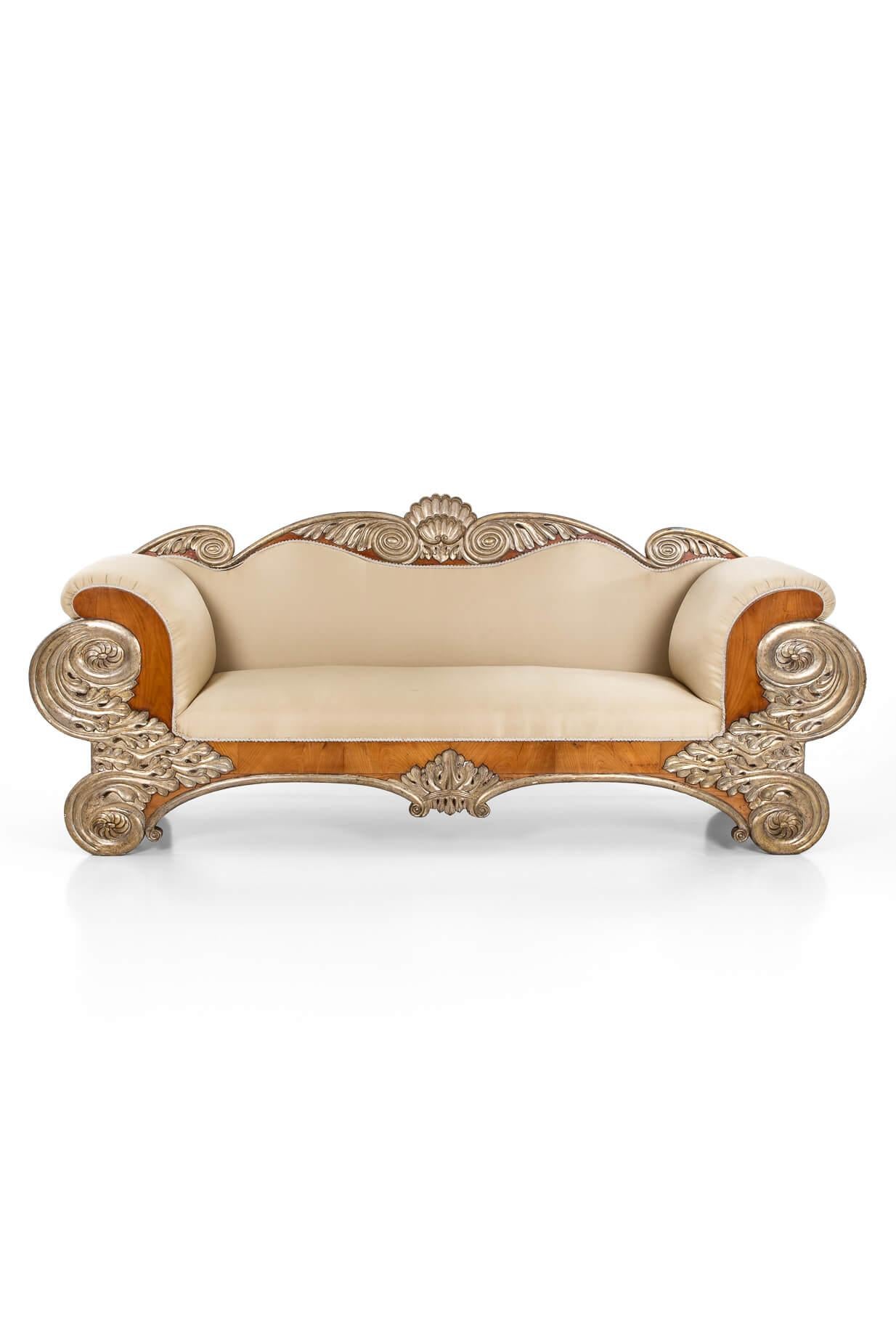 A commanding and elegant early 19th century Spanish salon sofa.

Carved in birch and giltwood, the generously padded back is crested with striking volutes, leafage and a central clam shell.

Each end of the scrolling arms is decorated with a gilded