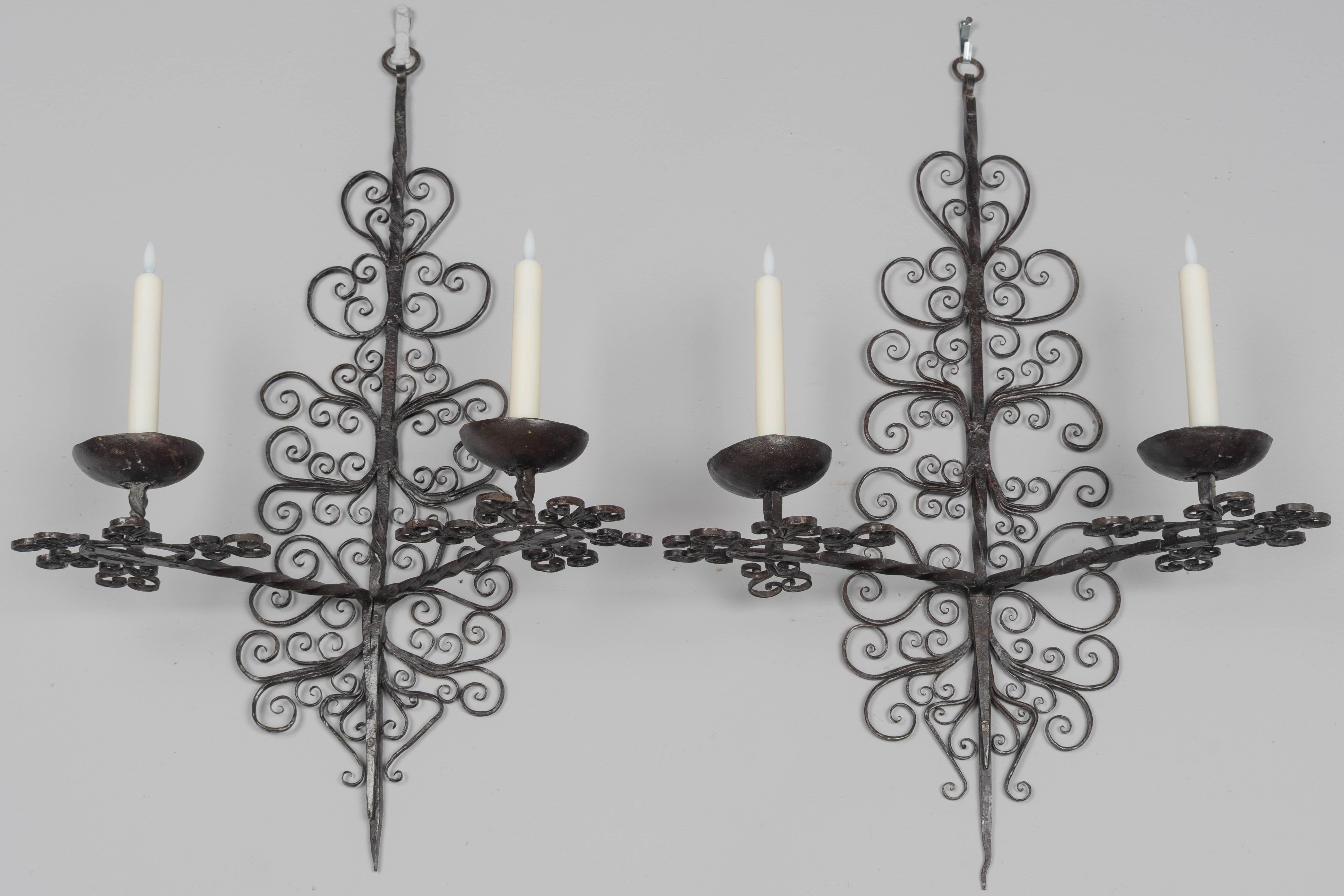 Coppia di appliques a due luci in ferro battuto in stile barocco spagnolo del XIX secolo con raffinata lavorazione a volute e coppe per candele martellate e rivettate. Ben dettagliato, realizzato a mano. Mostrato con candele a batteria, non incluse.
