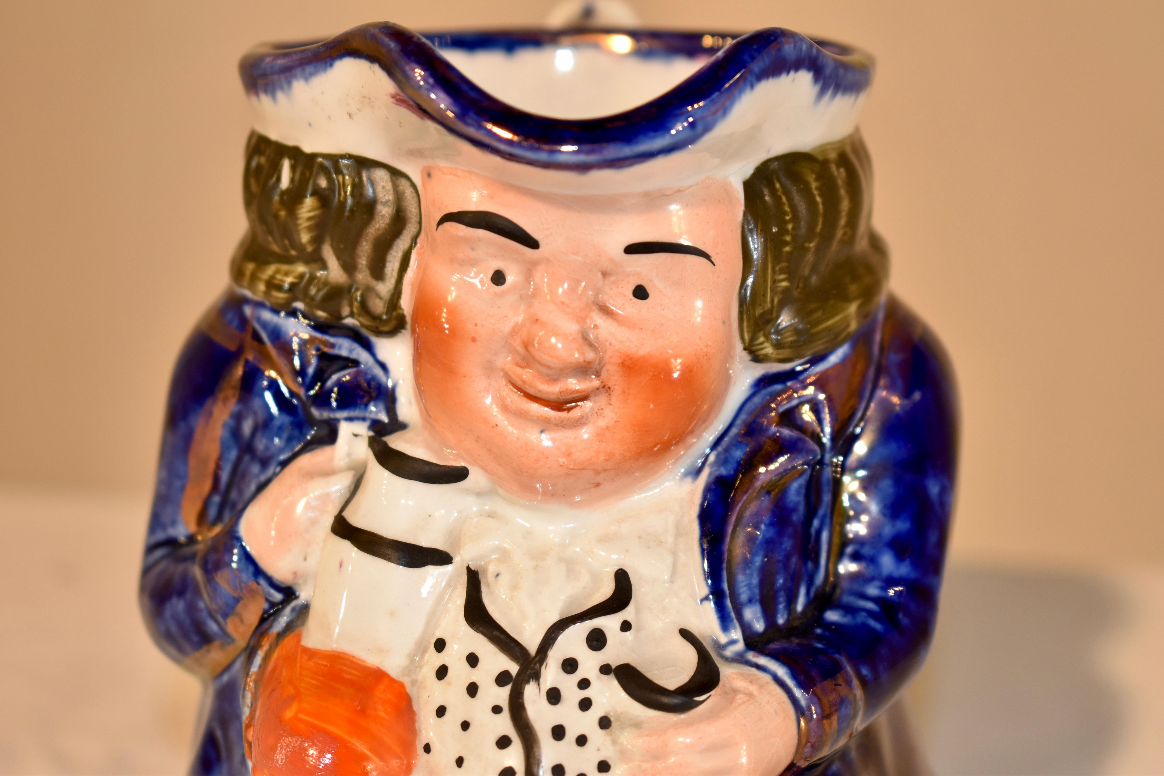 19th century ceramic Toby jug from the Staffordshire region of England.  He has a blue jacket and a warm expression on his face.  The jug has copper lustre accents as well for added definition.  