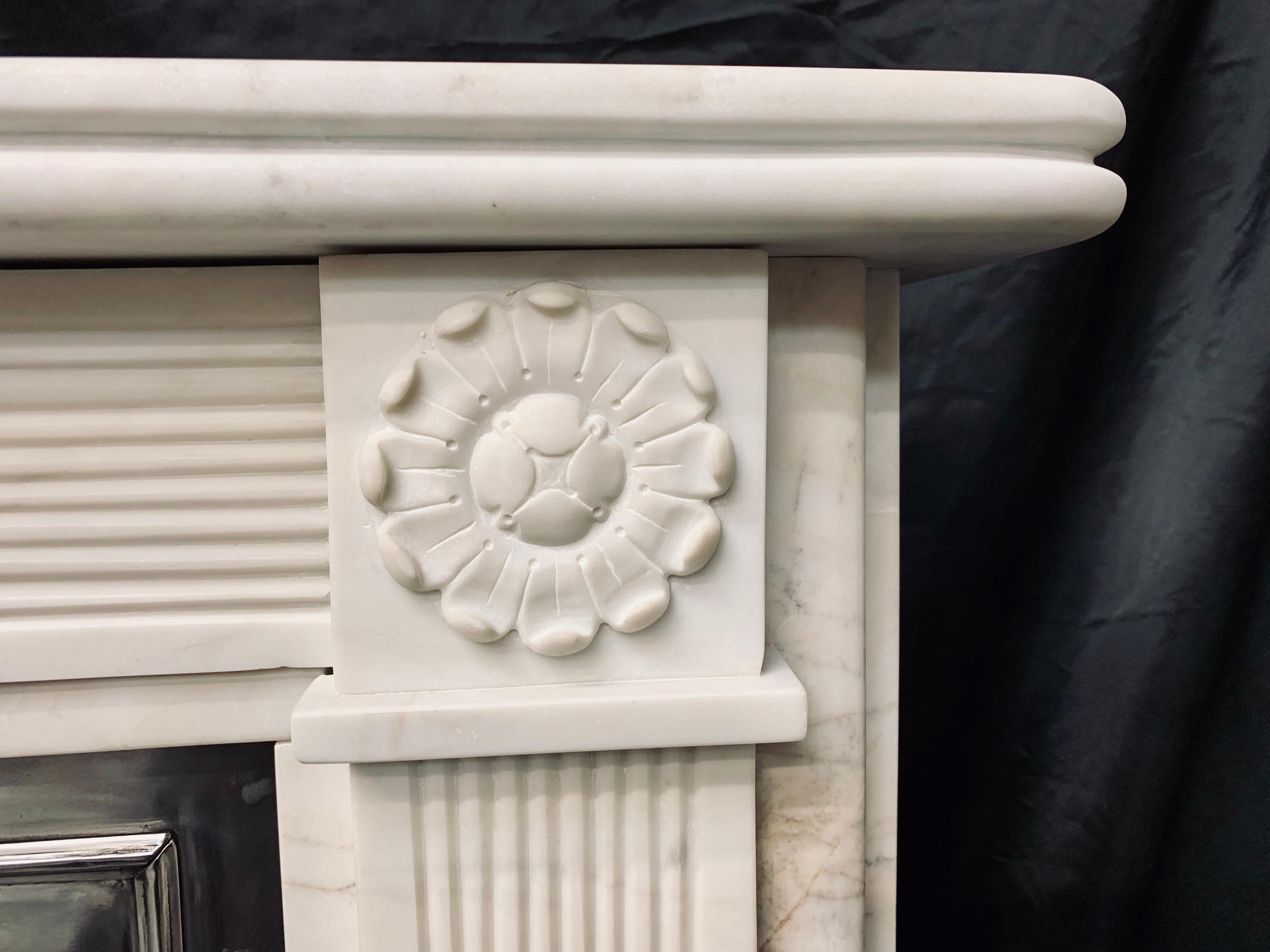 A small and attractive 19th century Regency style lightly veined statuary marble fireplace surround, a reeded shelf sits above a fine reeded stepped frieze, flanked by capitols with well carved rosettes supported by fine reeded jambs all supported