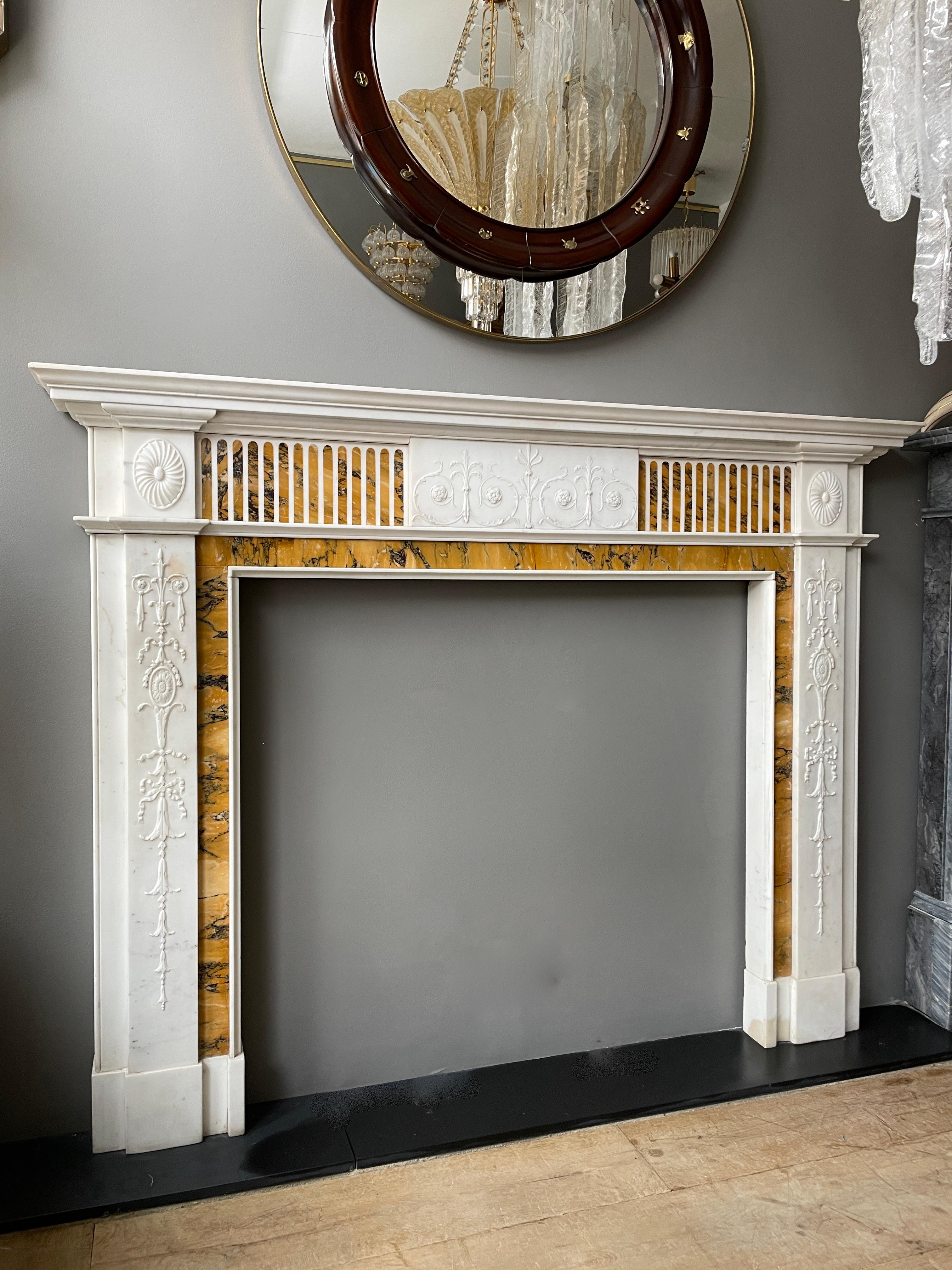 An elegant antique chimneypiece in Statuary White marble, and inlaid Convent Sienna. The jambs having delicately carved panels of ribbon tied bell drops, scrolls, foliage and oval Paterae. The conforming center tablet of scrolls, foliage and