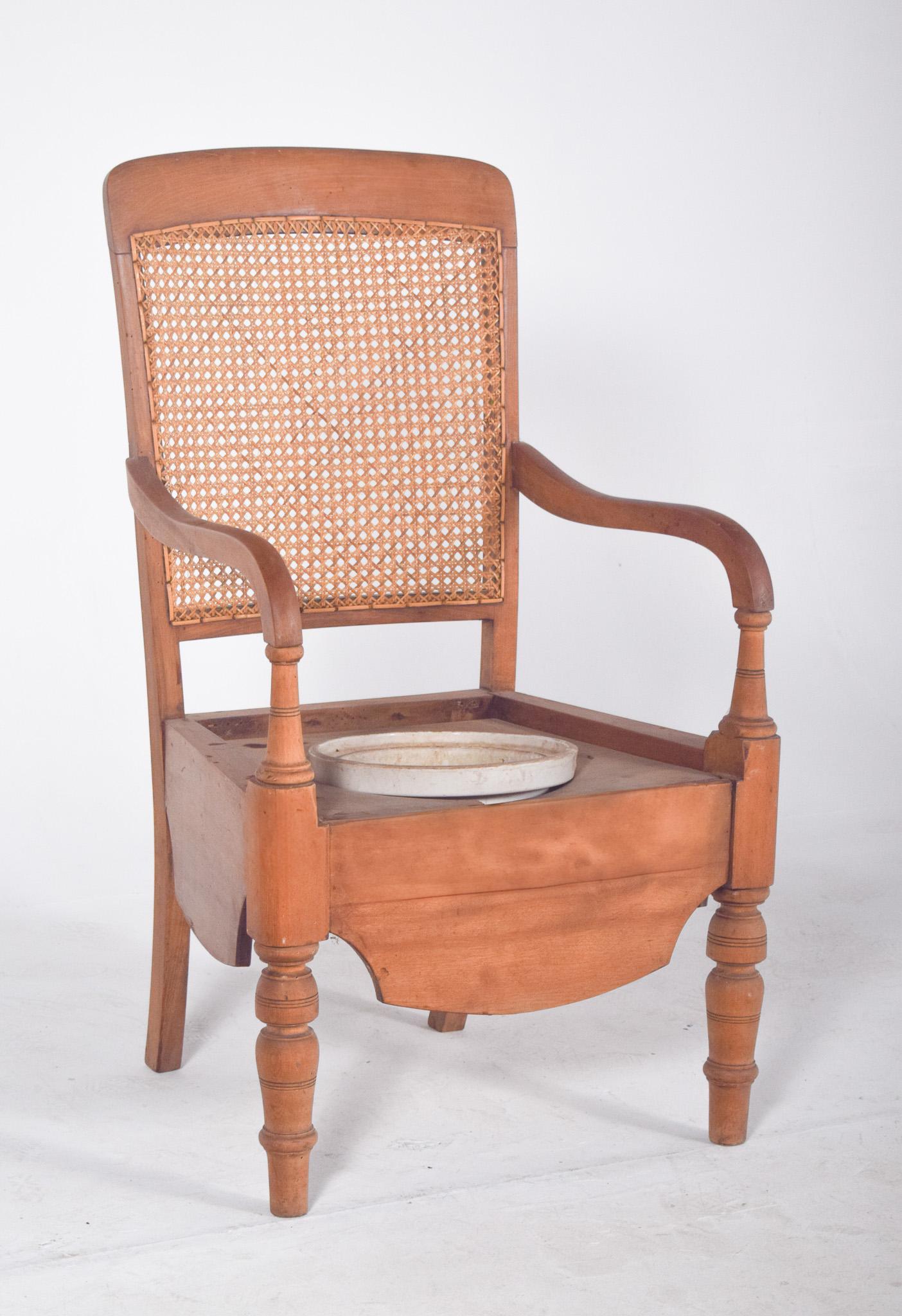 Chaise-pot avec dossier en paille. En bois clair, avec un siège creusé et une ouverture centrale où se trouvait à l'origine un récipient à vaisselle (pot). Les bras et les pieds tournés donnent à la pièce un aspect rustique qui mêle la simplicité à