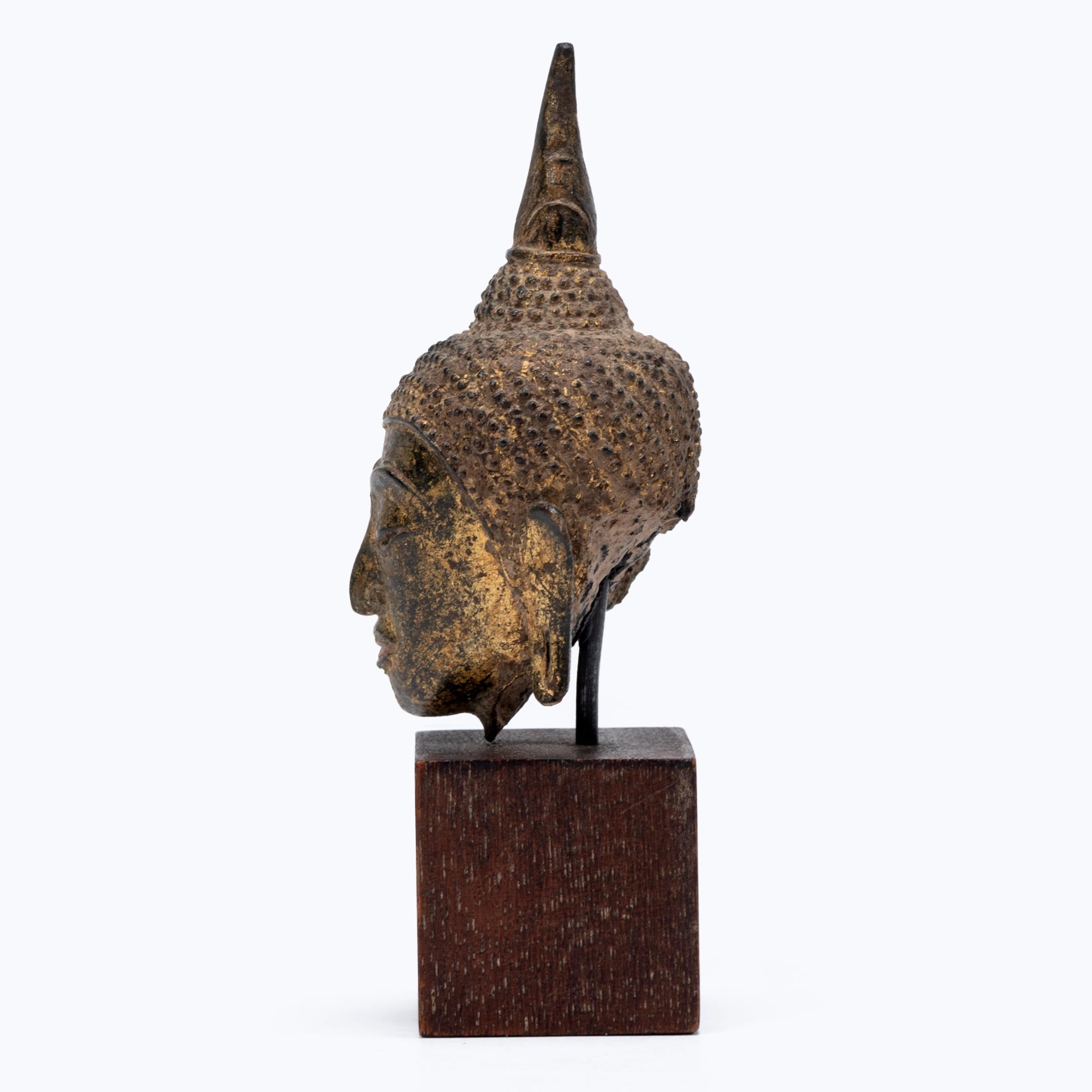 This serene Buddha head was cast of bronze in Thailand in the mid-19th century, and was originally ornately gilt and painted. Cast in the older Sukhothai style, the Buddha bears a flame-like spike stretching upward from the crown of his head. Called