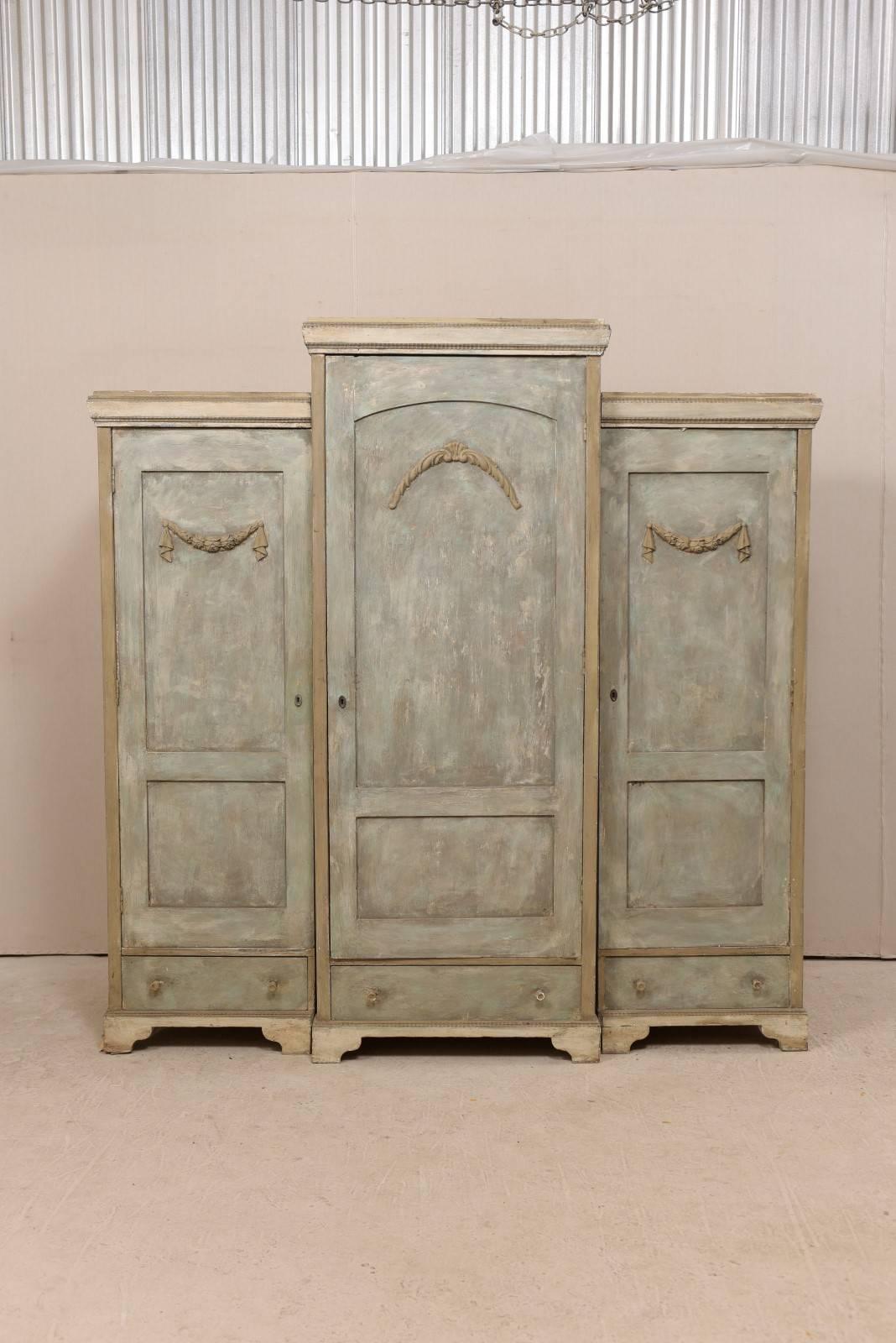 A Swedish breakfront three-door painted wood armoire from the 19th century. This antique Swedish storage cabinet features cabinets, each with a single door and bottom drawer. The three doors are each adorn with swagged appliques. The centre section