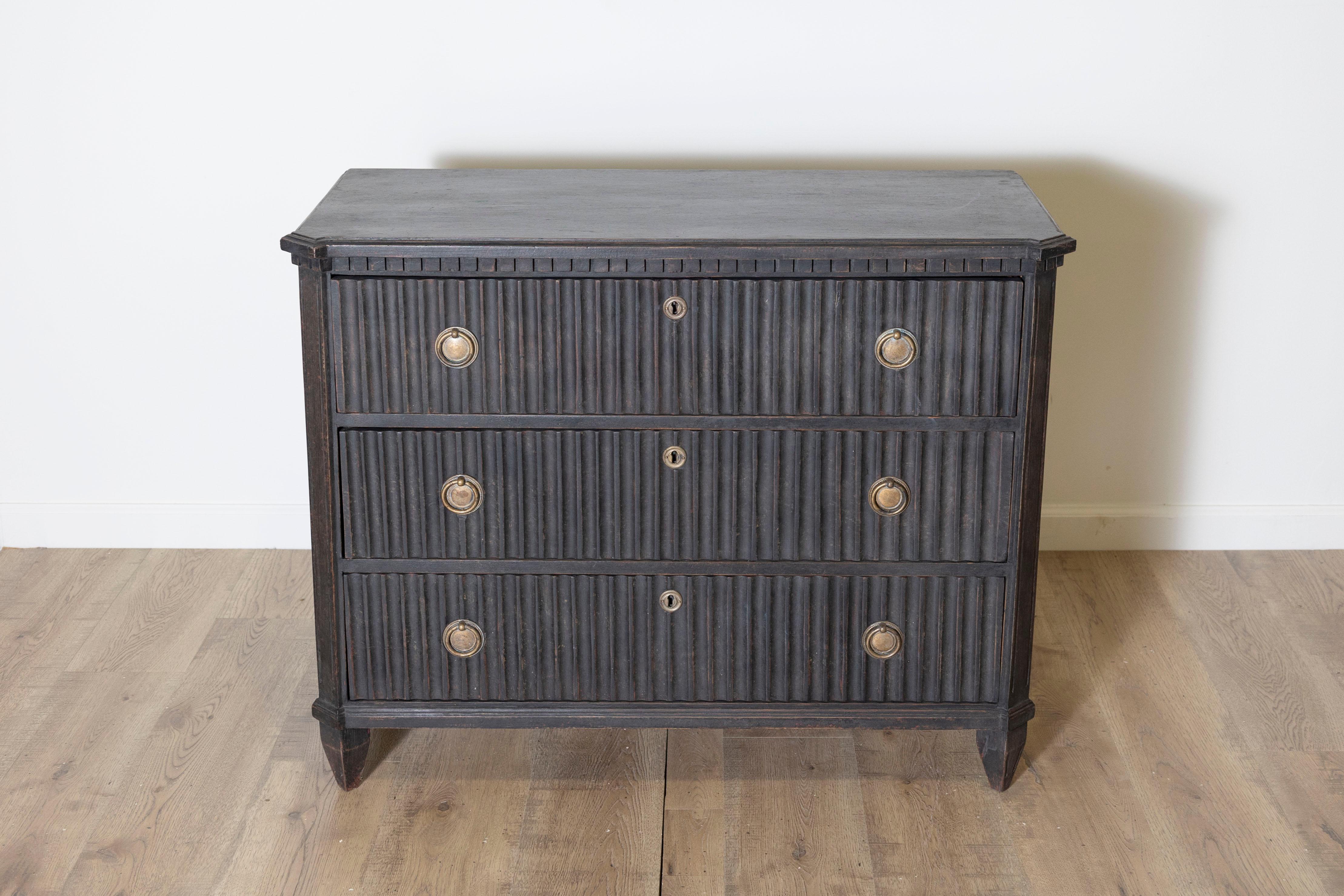 Questa cassettiera in stile gustaviano, risalente al 1860-1870 circa, mostra l'eleganza senza tempo dell'artigianato svedese. Rifinito in nero con una patina, il pezzo presenta una delicata striscia decorativa con denti sotto la parte superiore, che