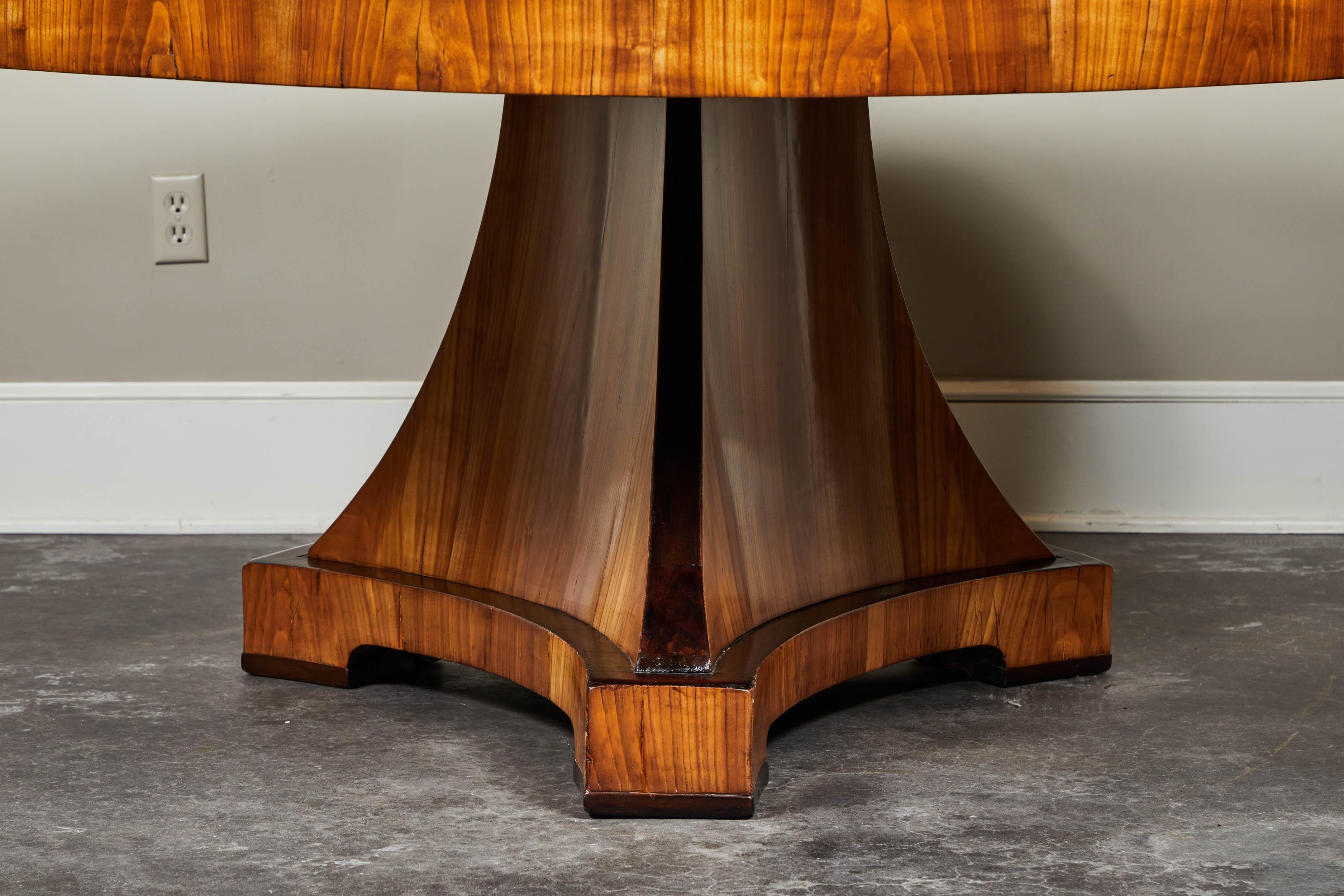 A round Biedermeier table, with a large group top on a tripartite base, with an elm veneer and ebonized trim detailing. This style of furniture, such as this lovely table, was meant to bring both elegance and utility to owners' private lives.
      