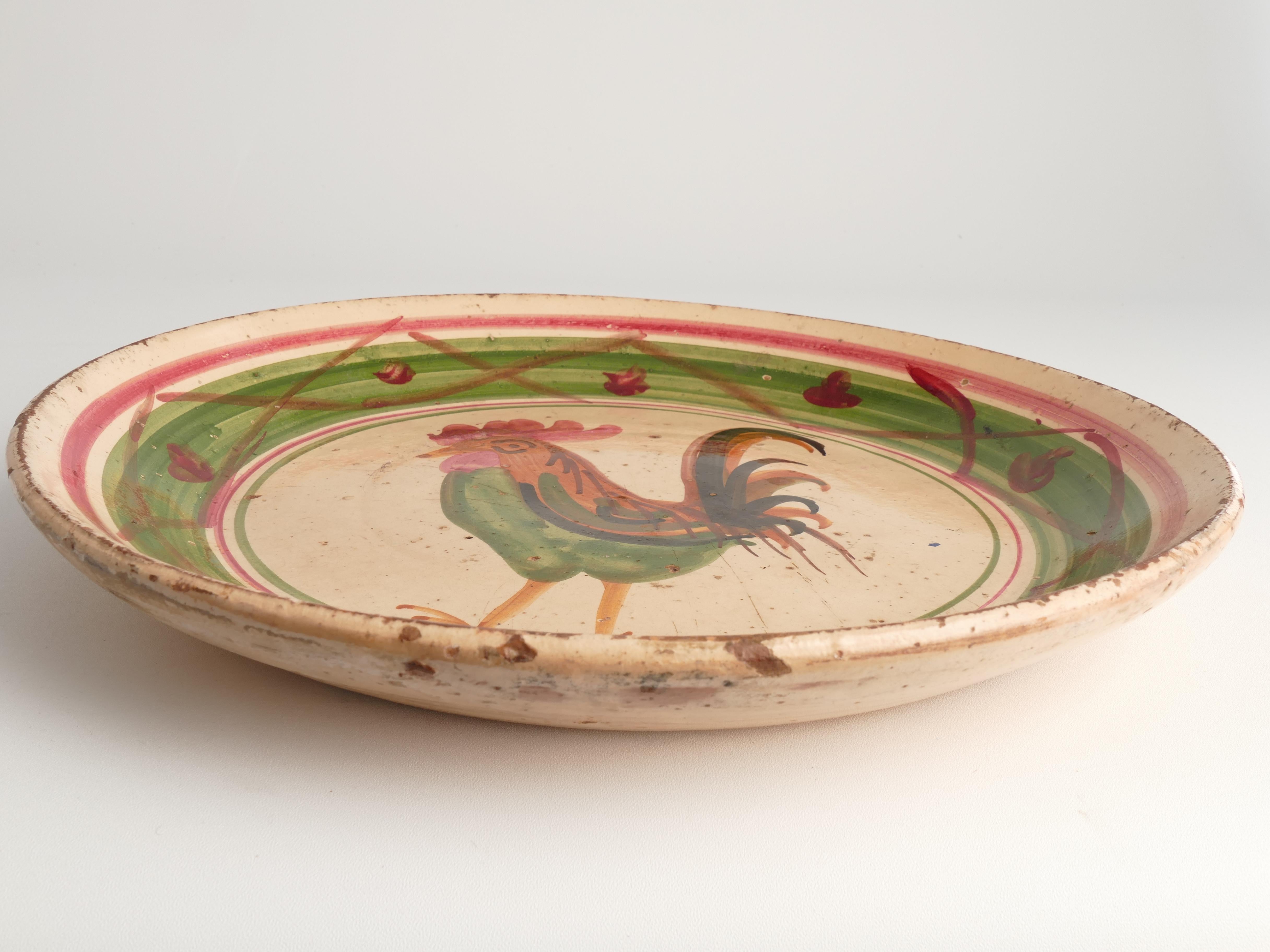 A large milk bowl crafted from ball clay features a rooster motif in red, green, and brown. This piece, originating from the mid-19th century, is adorned with engobe—a mixture of pipe clay and water, tinted with manganese for brown, copper oxide for