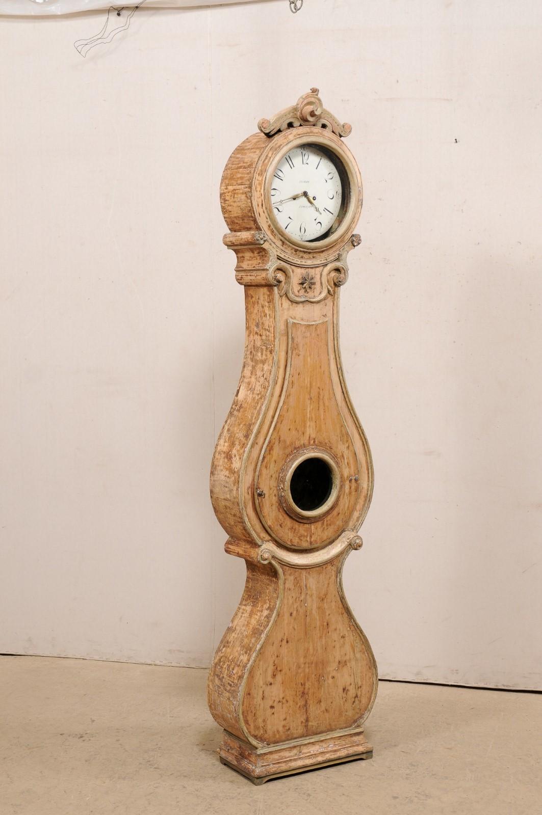 A Swedish Fryksdahl tall floor clock from the 19th century. This antique Fryksdahl clock from Sweden features a nicely carved bonnet top, petite flowers, volute accents, and a central star adorn the neck. The tear drop shaped belly has a glass