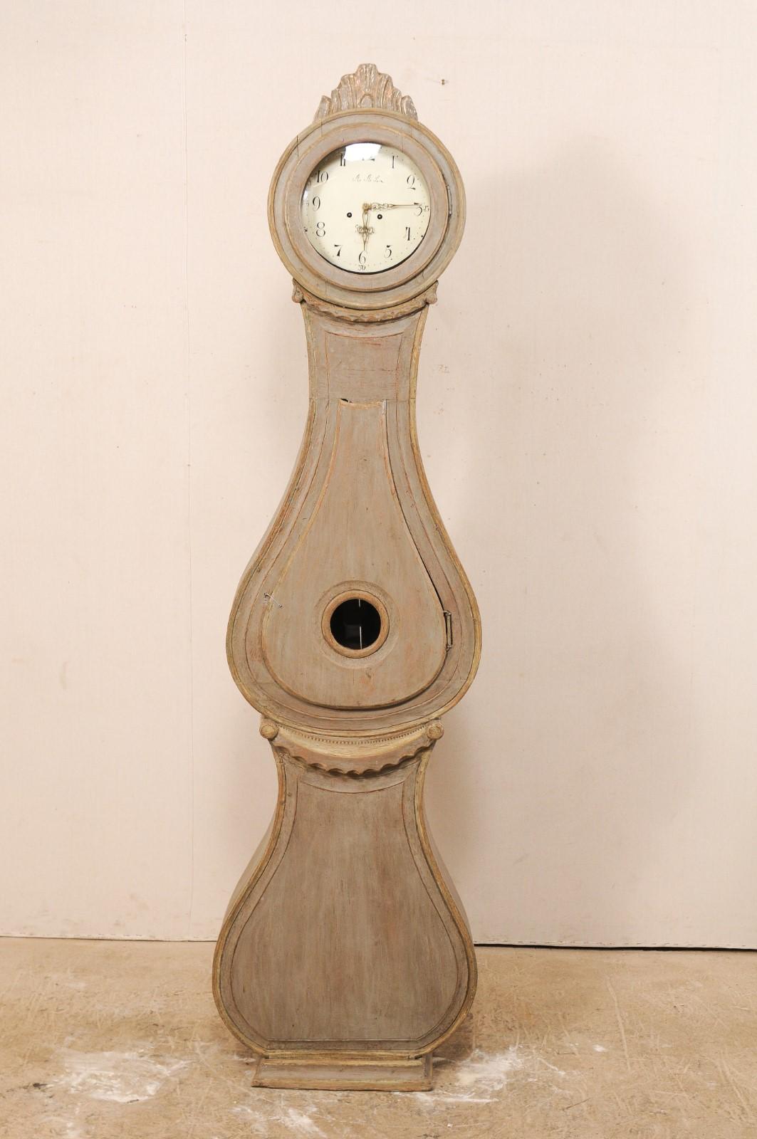 A 19th century Swedish painted wood Fryksdahl clock. This Fryksdahl clock features a carved crest of fanned foliage, scalloped carved swags about the neck and waist flanked with volute accents, a rounded head and curvaceous belly and base. This