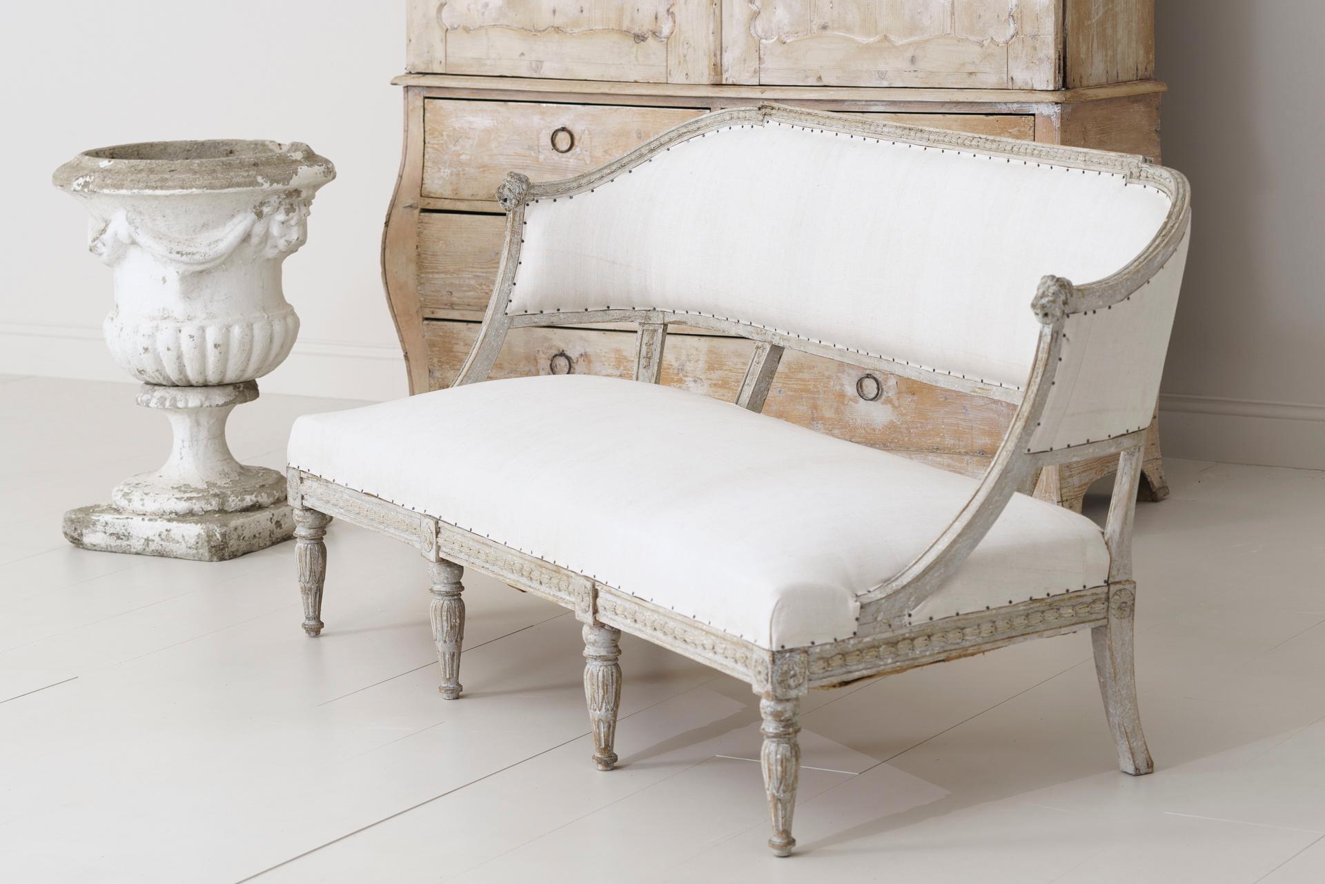 A 19th century Swedish barrel back sofa settee in the Gustavian style, newly upholstered in antique linen, circa 1850. Hand scraped to reveal the original paint. The frame features carved lion heads and bell flowers while the front legs are adorned