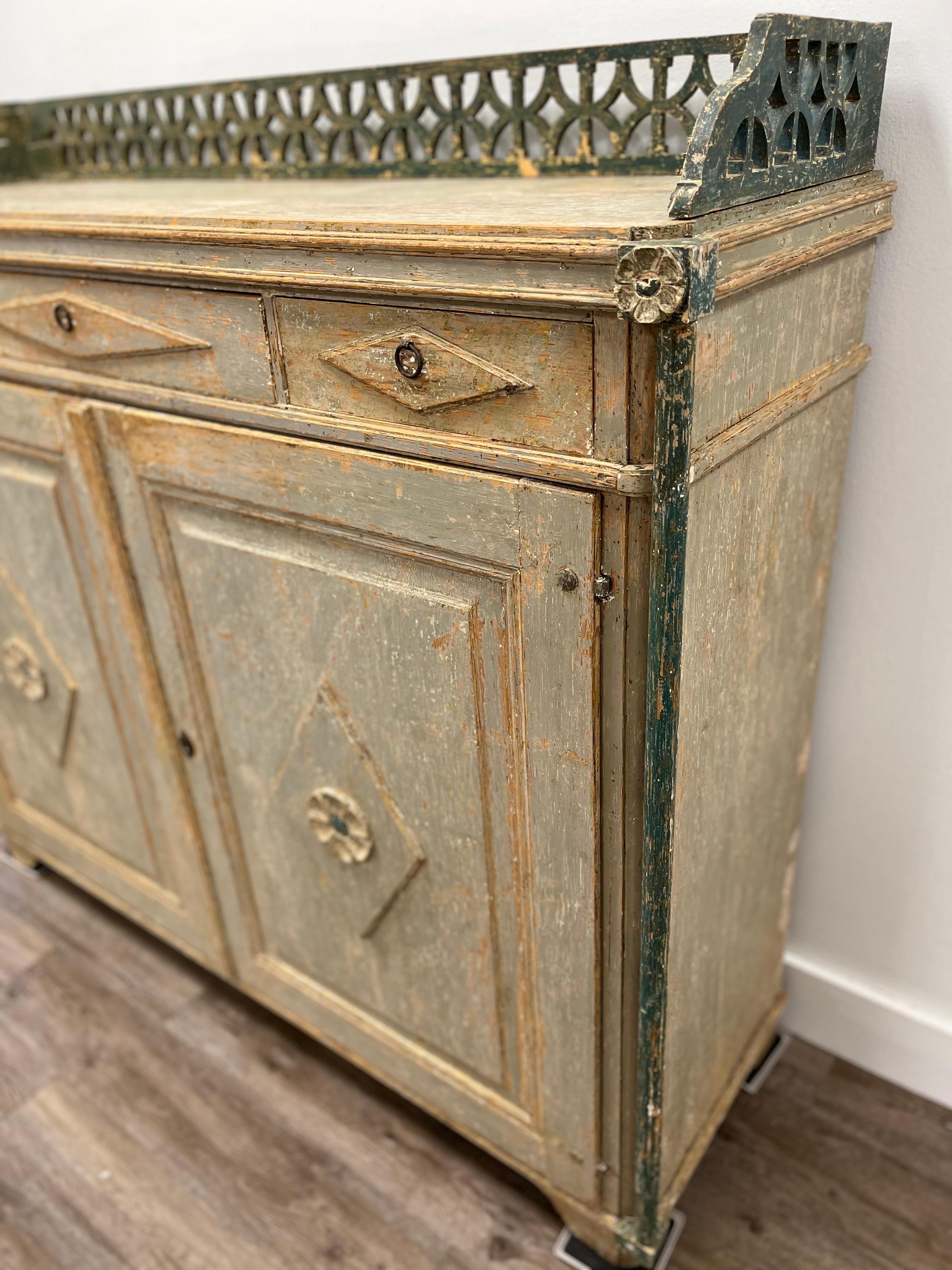 A Swedish Late Gustavian buffet from Medalpad. Detailed wood carvings – flower petal décor and pierced gallery top. Three drawers with horizontal diamond-shaped fronts and simple pulls on top. Below, recessed cabinet door fronts with vertical