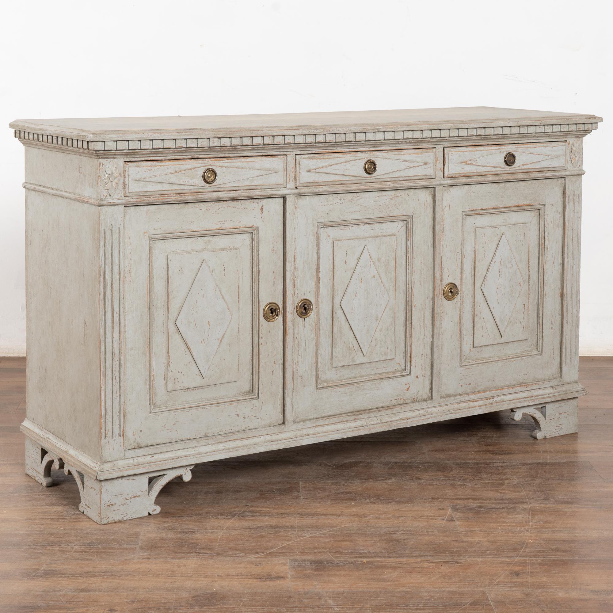 Swedish Gustavian gray painted pine sideboard or console with upper dentil molding and one interior shelf.
Traditional diamonds accent the three drawers with brass pulls over three doors with keys and locks which function stiffly, buyer may opt to