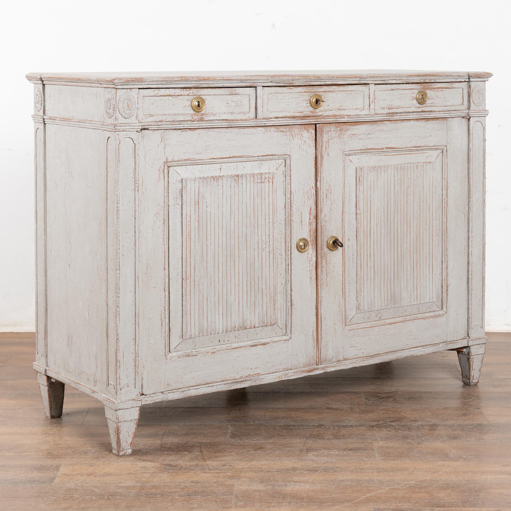 Questa credenza svedese in stile gustaviano ha una finitura dipinta di un grigio tenue che completa il tradizionale intaglio scanalato lungo le ante a pannelli e i piedi affusolati.
I tre cassetti funzionanti si aprono con una chiave scheletrica (1