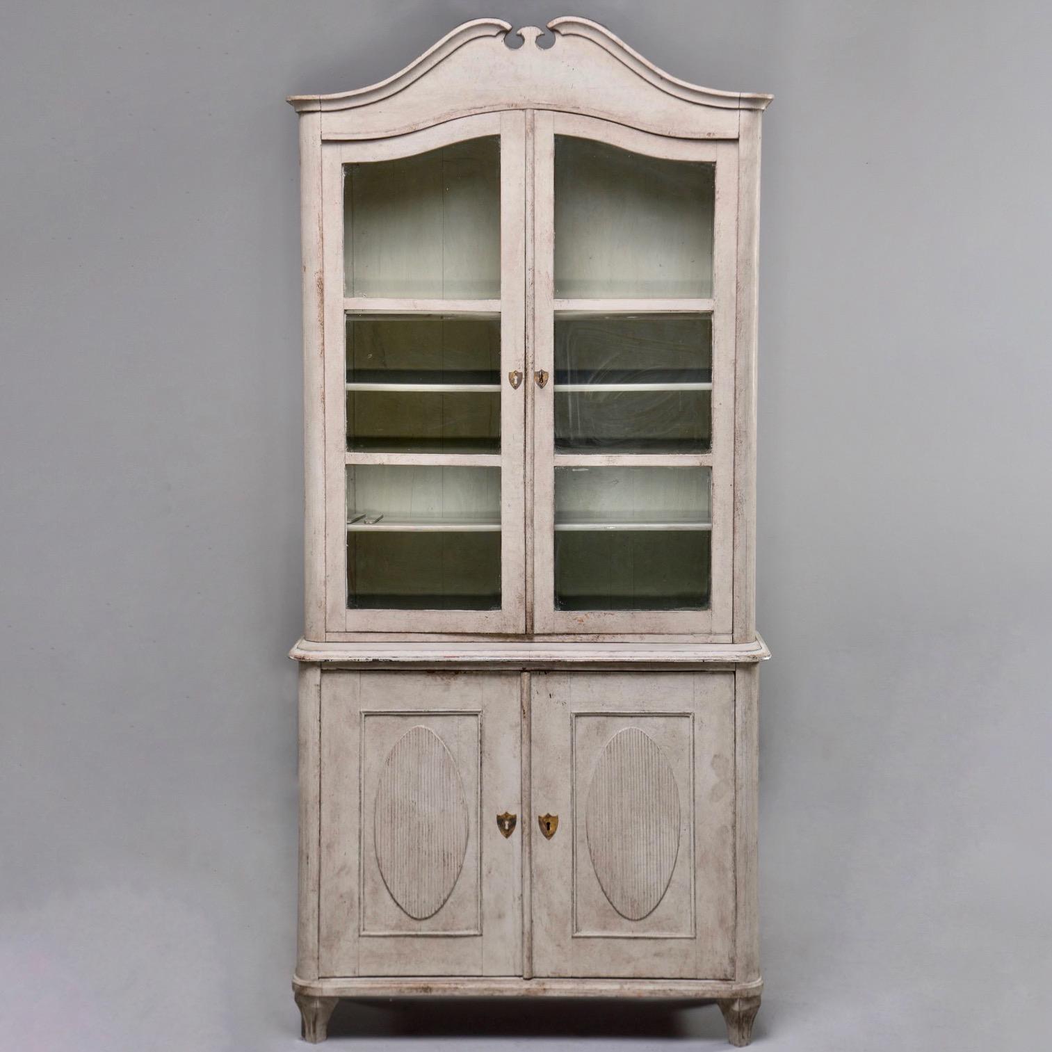 Swedish tall pine Gustavian style cupboard has greige painted finish and glass front doors on the top section with a functional lock and skeleton key, circa 1860s. Top portion has three internal adjustable shelves. Bottom section has decorative