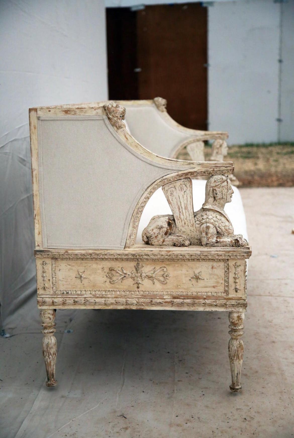 An absolute stunning piece of artisan craftsmanship here. The gorgeous 19th century Swedish sofa features Sphinx and Lion motifs with a cream color palette. With significant size and presence, this armed bench has a long single seat with curved arm