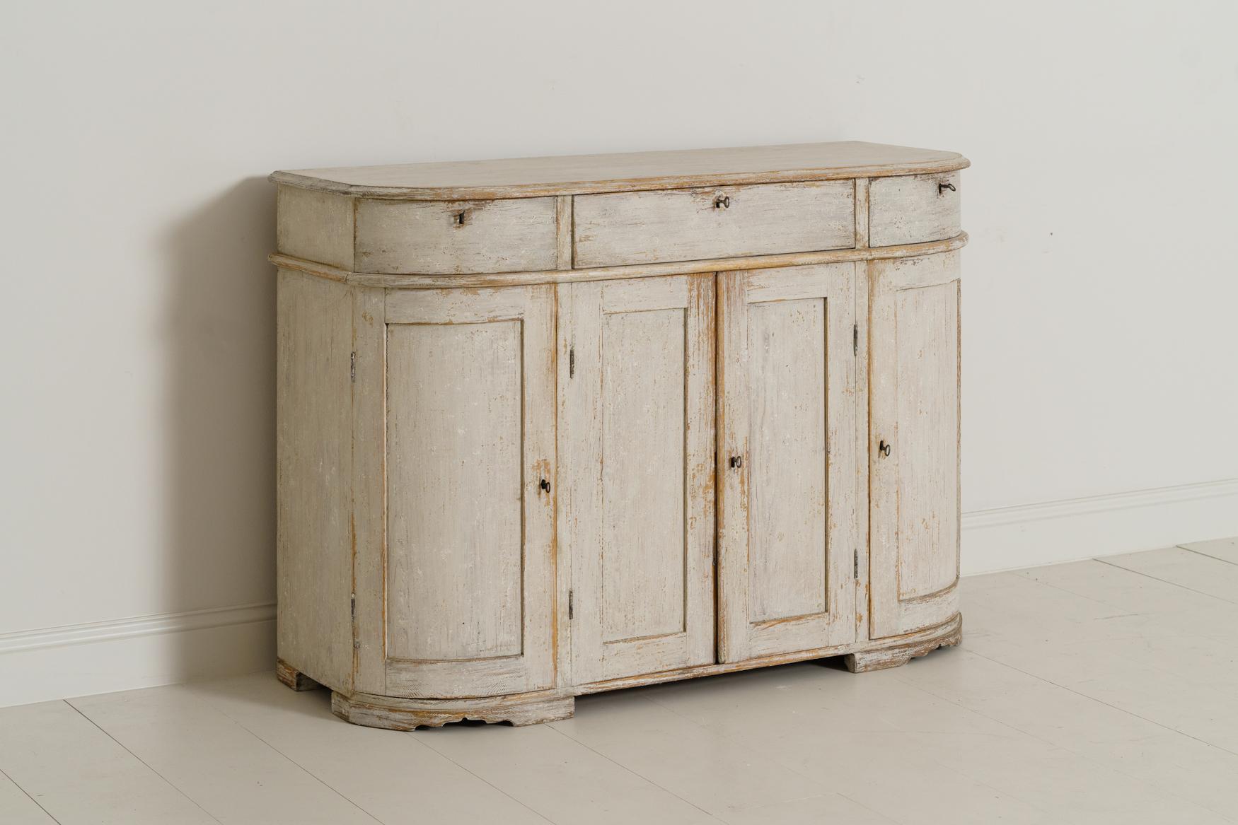 A Swedish demilune sideboard or server from the 19th century in the Gustavian style. The patina is a beautiful chalky old white with ivory undertones and wood showing through in areas. The buffet has three drawers and four doors. The centre section