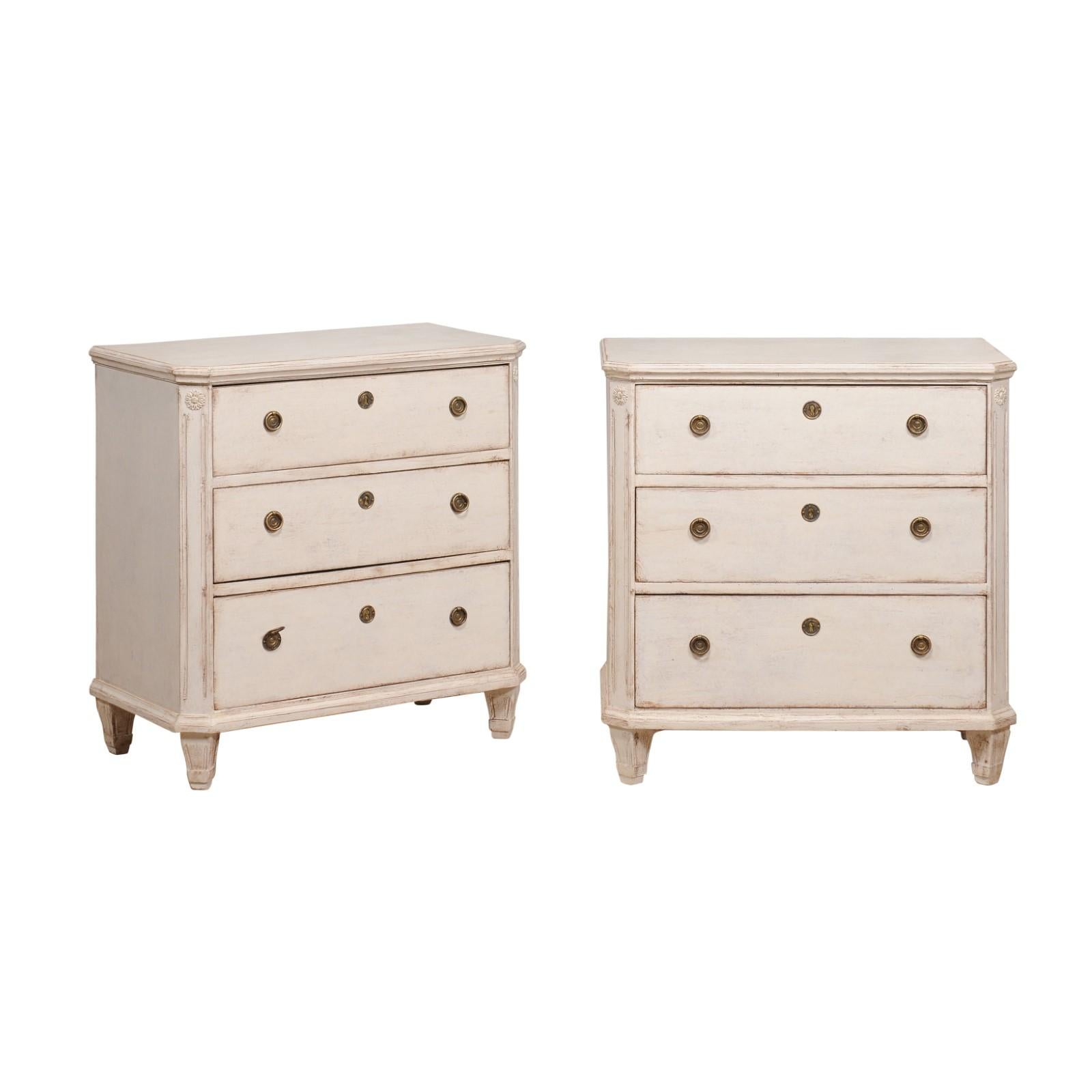 A pair of Swedish Gustavian style chests from the 19th century with off white / beige painted finish, three drawers, carved rosettes and tapered feet. Immerse yourself in the effortless grandeur of this 19th-century pair of Swedish Gustavian style