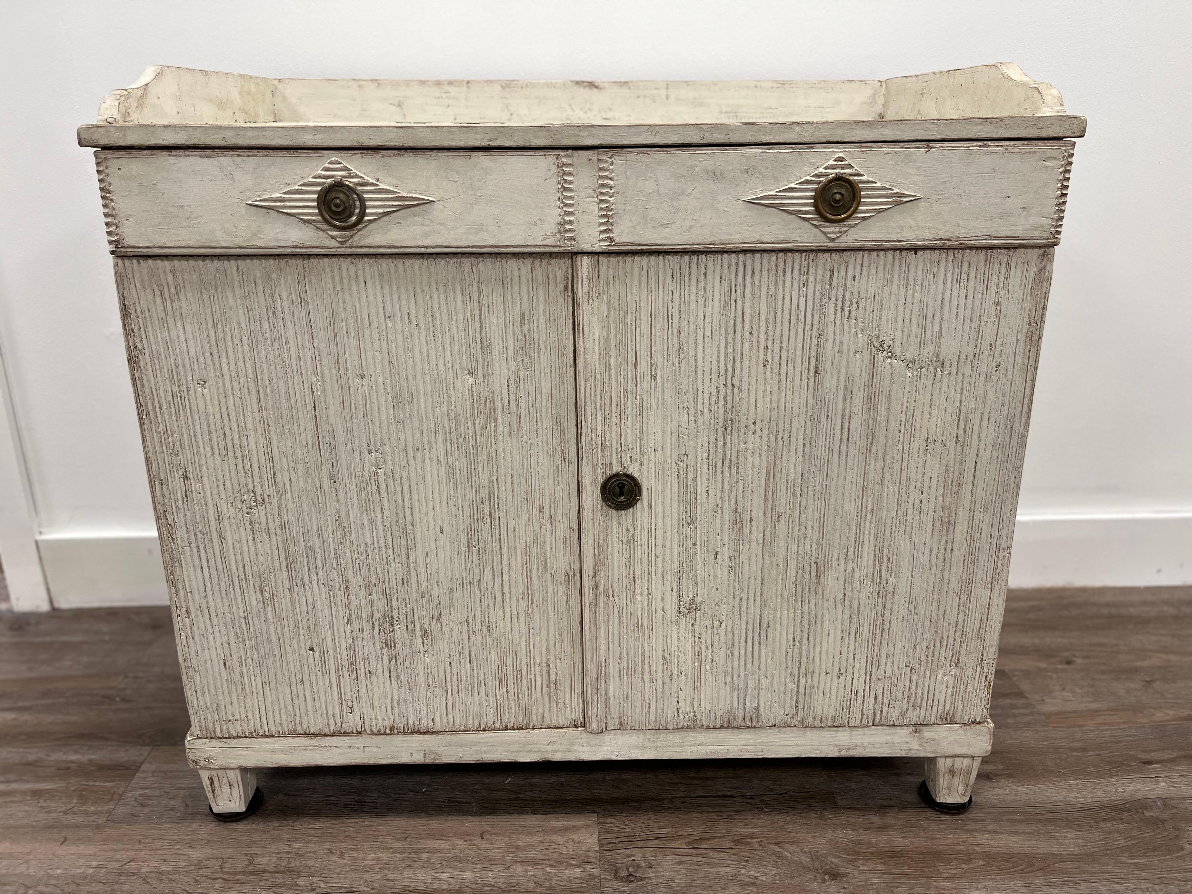 A small provincial Gustavian Style sideboard with original hardware and repainted in light grey. One wide drawer with two reeded diamond-shaped details on the pulls. Sits atop two vertically reeded cabinet doors. Inside, two shelves.