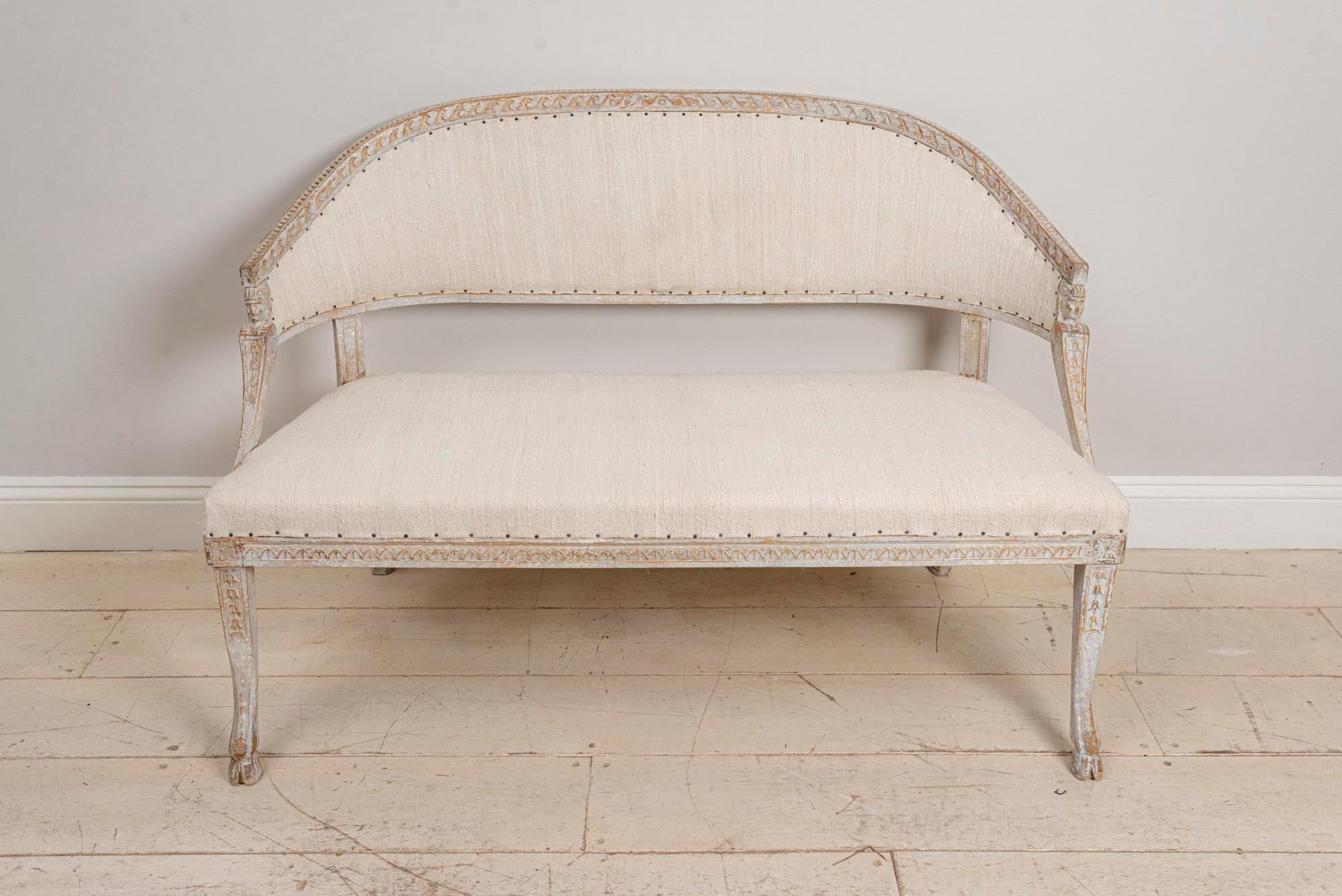 Early circa 19th century Gustavian two-seat Swedish sofa featuring a barrel curved back, Egyptian decorative detail, splayed legs and goat's hoof feet. The paint has been refreshed and has also been newly upholstered in a neutral linen. This lovely