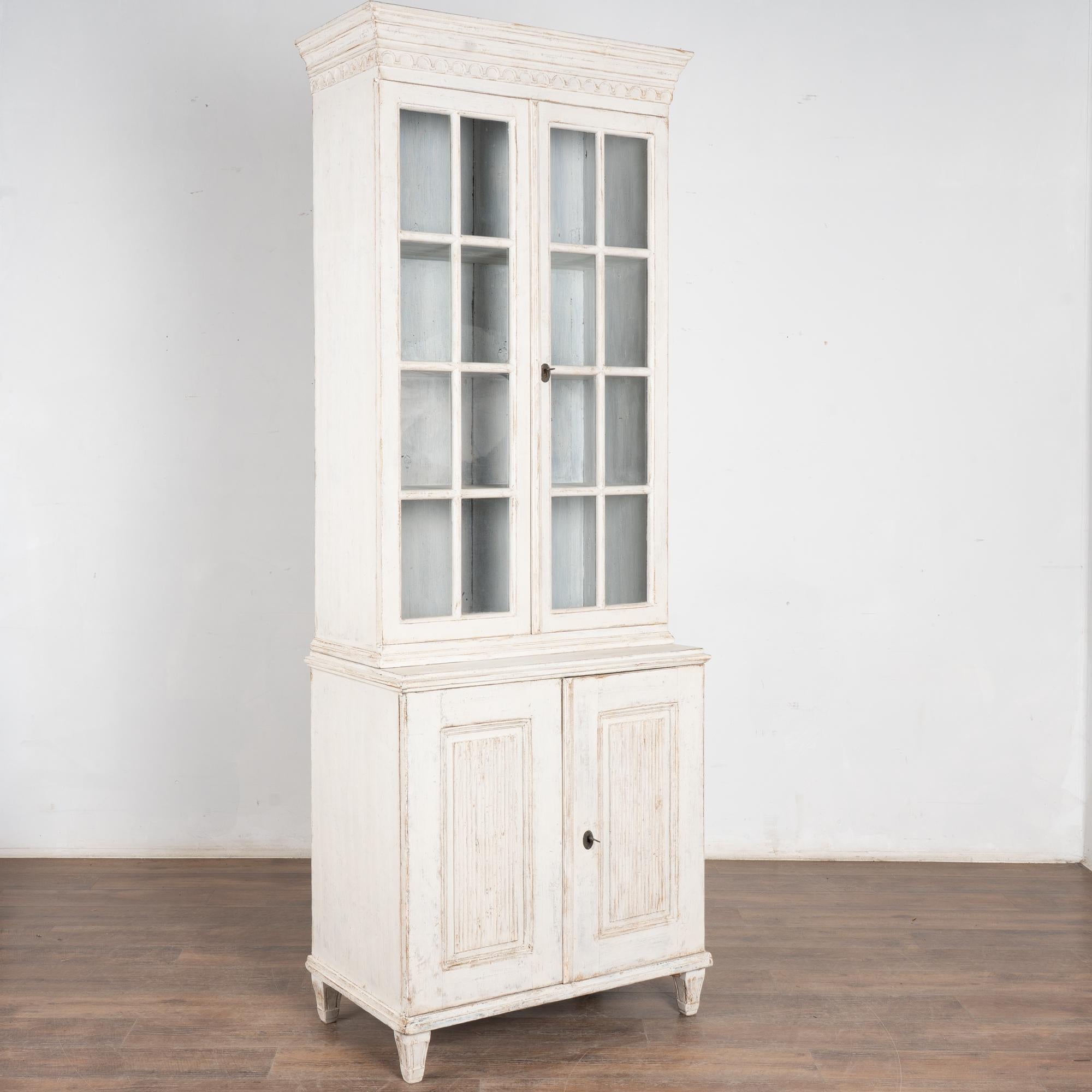 Questa stretta vetrina svedese può essere utilizzata anche come libreria. Le ante a pannelli scanalati e l'intaglio decorativo lungo la corona conferiscono a questo mobile bianco un fascino gustaviano.
Le ante superiori in vetro consentono di