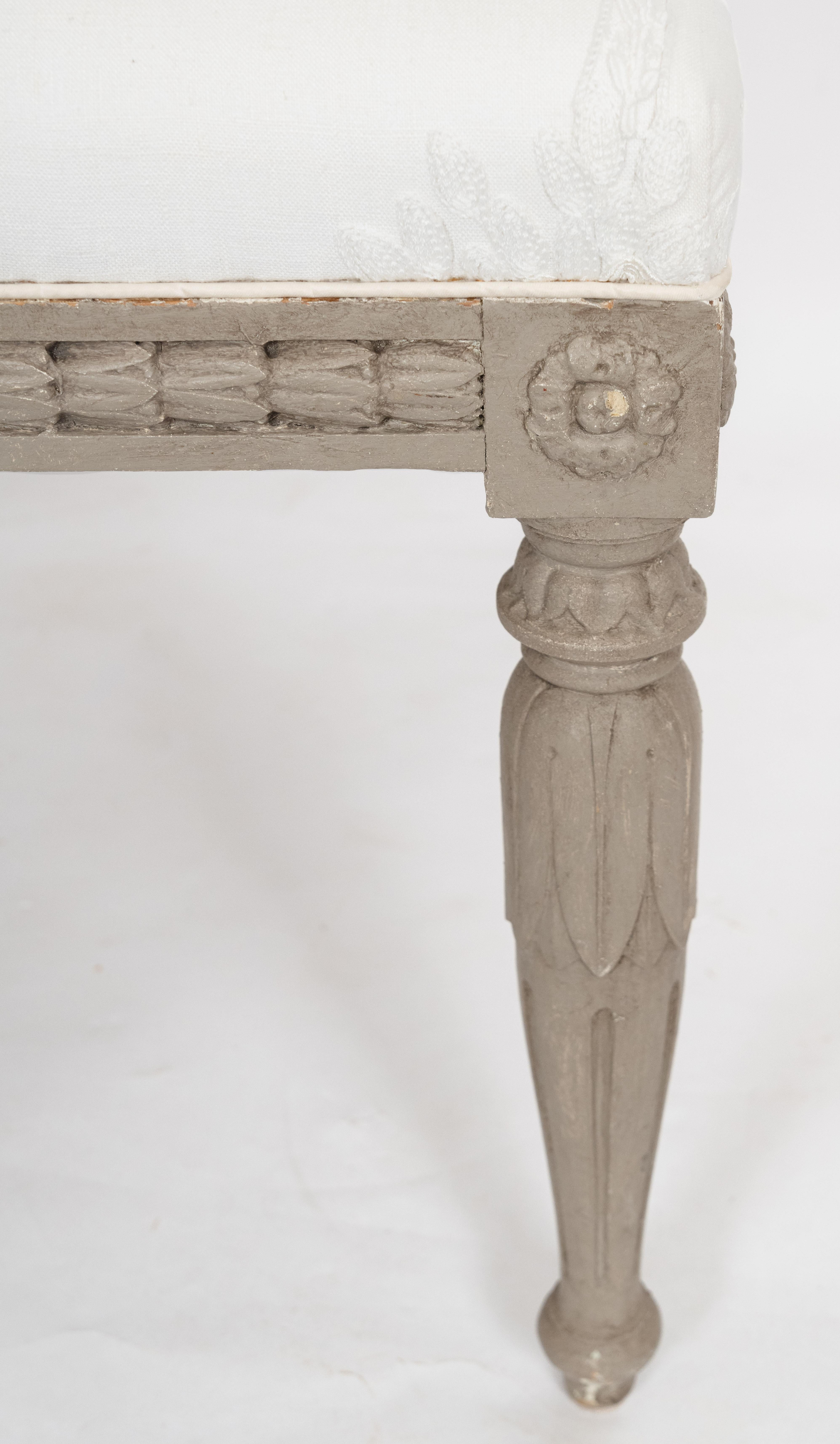Gustavianischer Fußschemel aus dem 19. Jahrhundert mit kunstvollen Schnitzereien entlang des Holzsockels. Die Umrahmung mit eingesetztem Glockenblumenfries wird von vier konisch zulaufenden Beinen in Form von palmettenförmigen, kannelierten Säulen
