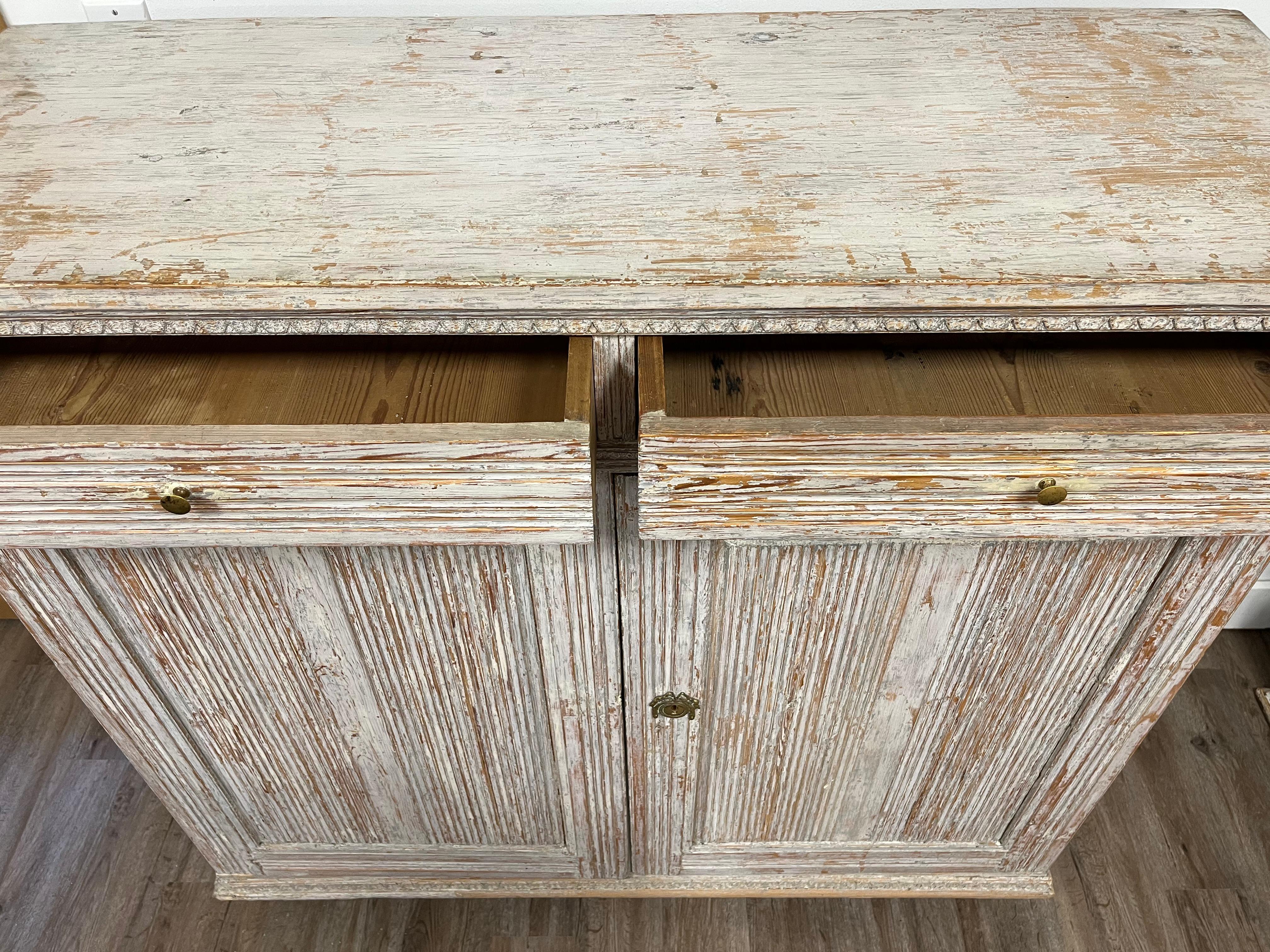 A large Swedish Late Gustavian buffet with elaborate wood carvings – bands of leaf cut detail surround the horizontal reeded drawer fronts and continue along the sides. Additional reeding frame inset vertical reeded door fronts. Front opens to