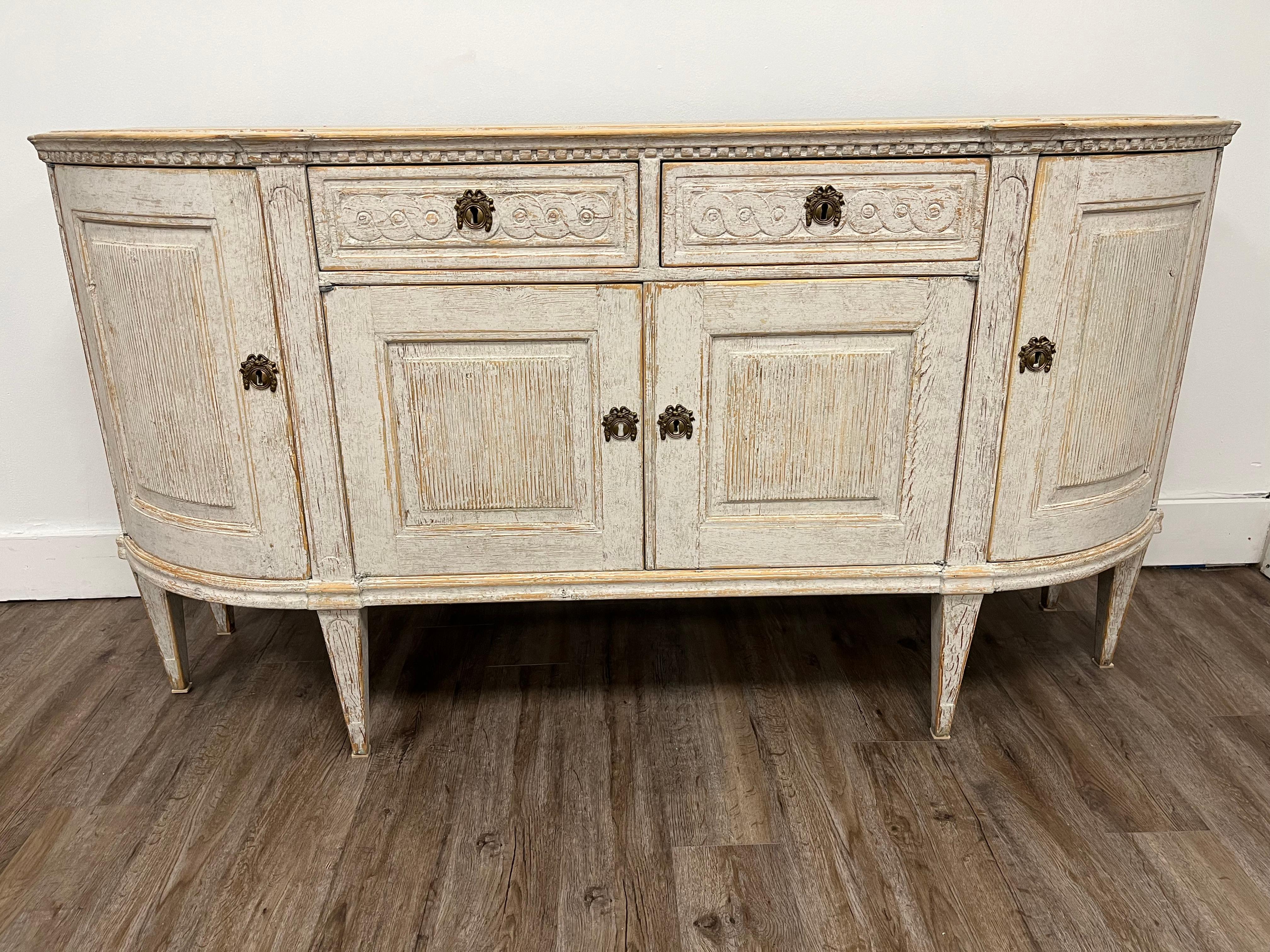 A Swedish late Gustavian sideboard with original brass hardware and locks. Tastefully repainted in pale grey. Featuring curved corner design, beveled top, decorative hand carved dental detail, Guilloché scrolling drawer front motif and vertical