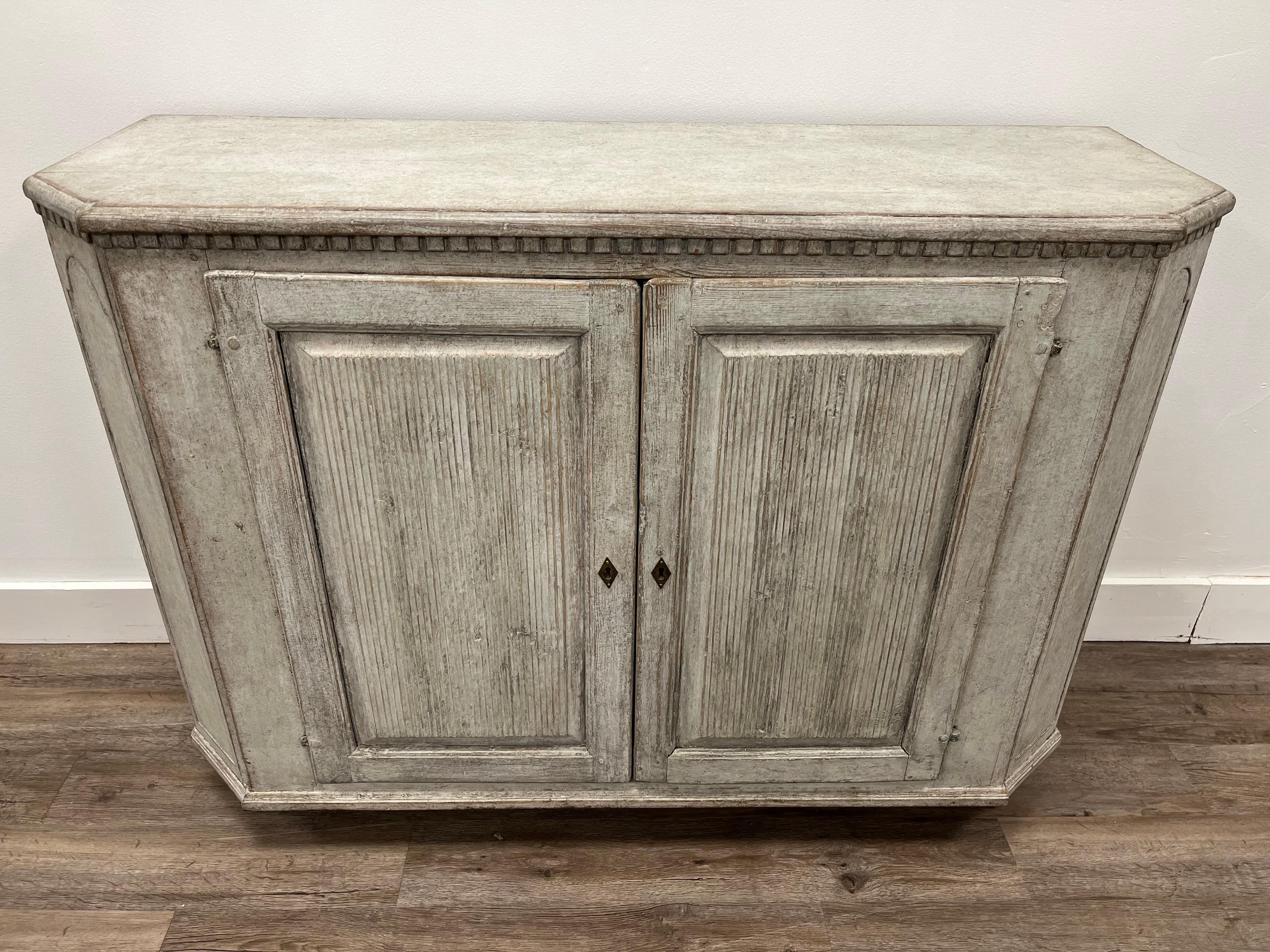A provincial Late Gustavian sideboard with dental frieze top and canted corners with recessed design. Vertically reeded door fronts open to two large shelves. Sits atop small tapered feet. Later grey exterior and soft yellow interior paint. Original