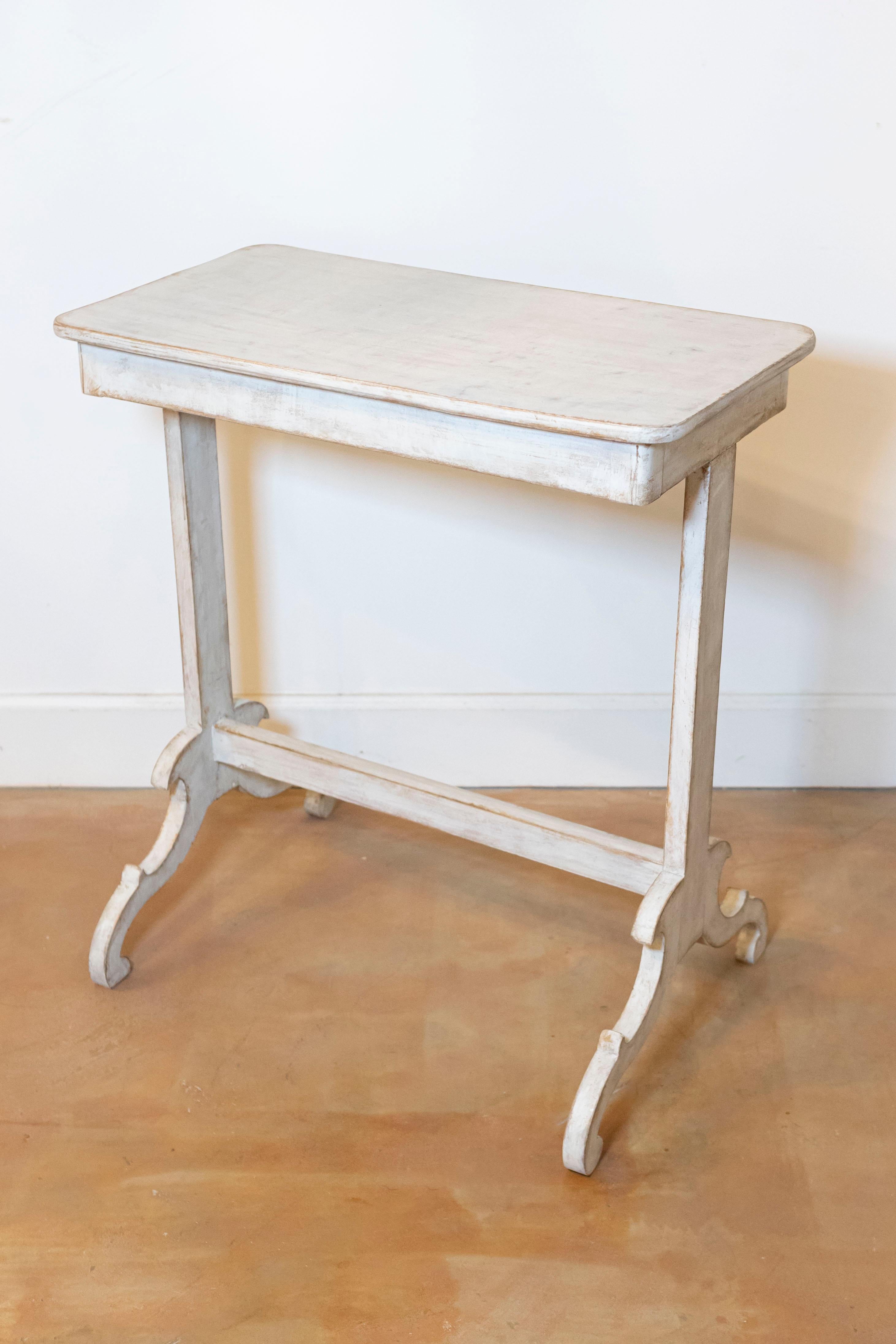 A Swedish side table from the 19th century with light gray painted finish and carved trestle base. Unveil the understated elegance of this 19th-century Swedish side table, a genuine antique treasure that whispers tales of yore. Coated in a serene