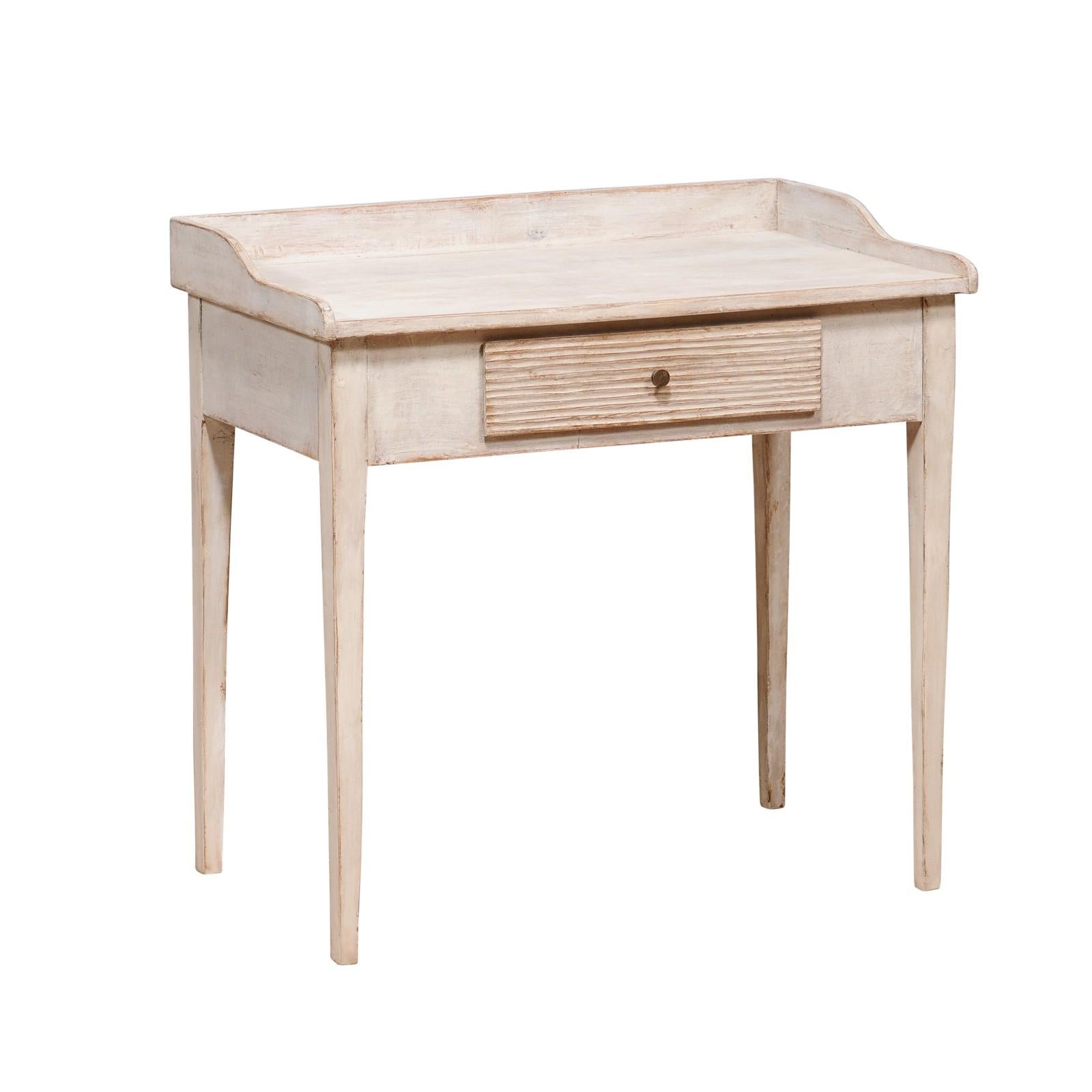 A Swedish off-white, light grey painted writing desk from the 19th century with three-quarter gallery surrounding the rectangular top, single drawer carved with reeded motifs and four tapered legs. Dive into the timeless charm of Scandinavian
