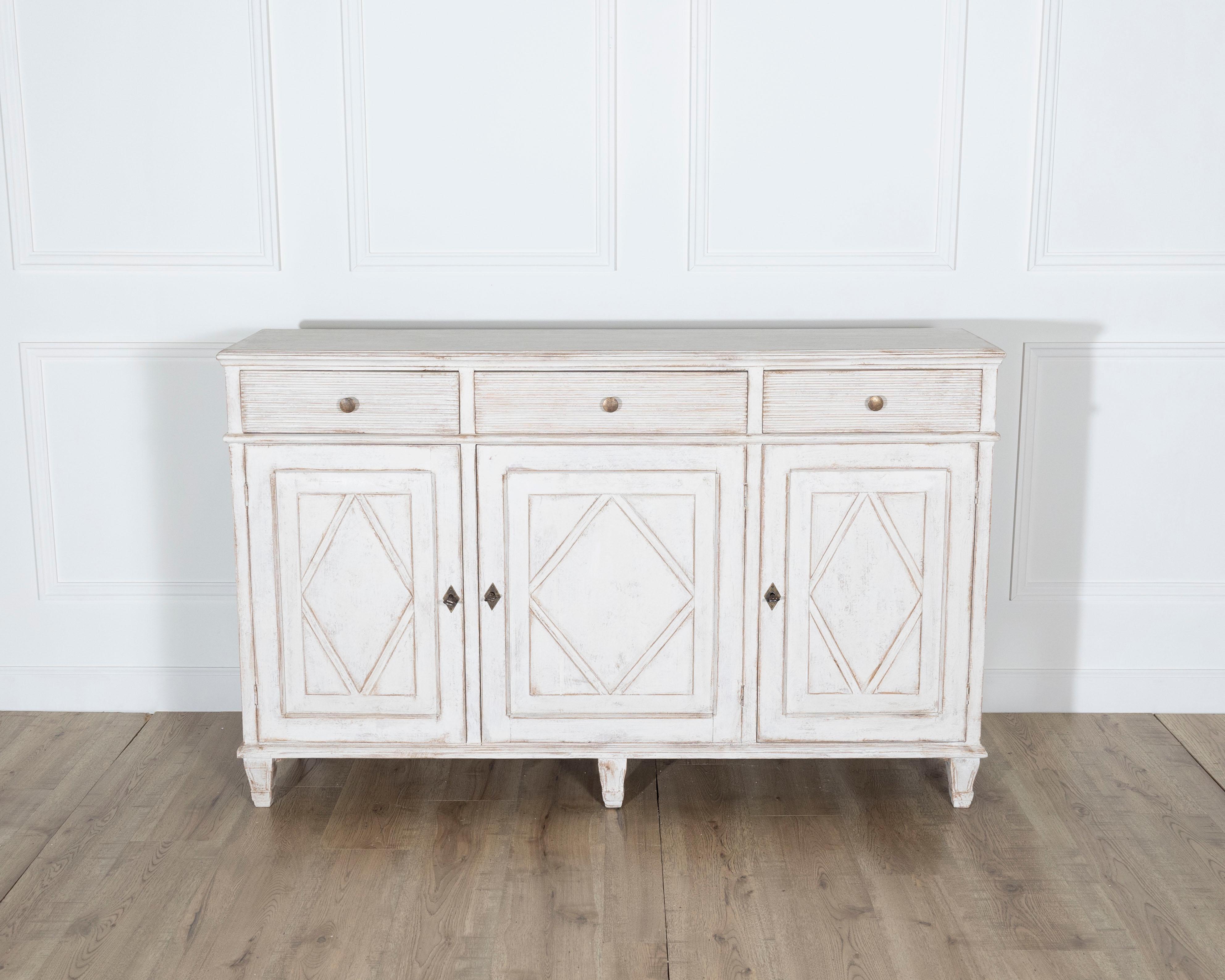 A graceful 19th century Swedish sideboard crafted from solid pine and finished in a soft, pale Gustavian-inspired paint with gentle distressing that enhances its age and authenticity. The piece features three drawers across the top, each with simple