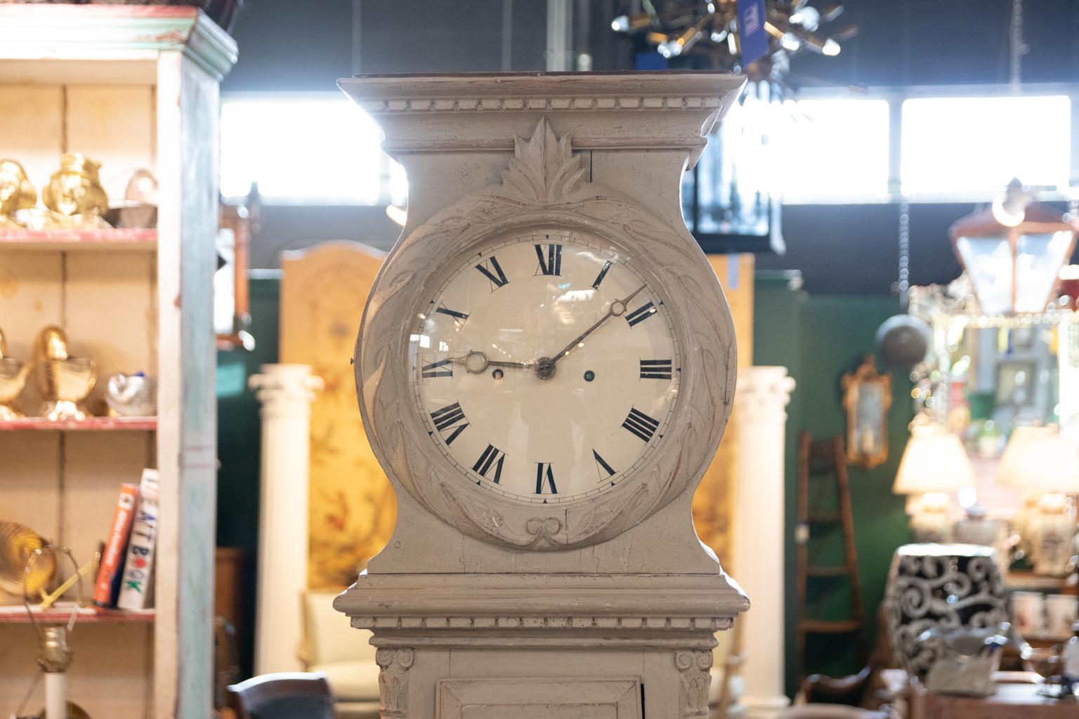 Swedish Gustavian style painted tall case clock in working condition, circa 19th century. The case is detailed with dentil trim, carved folliage framing the face, and composite columns on the body. The piece chimes every hour. Please note of wear
