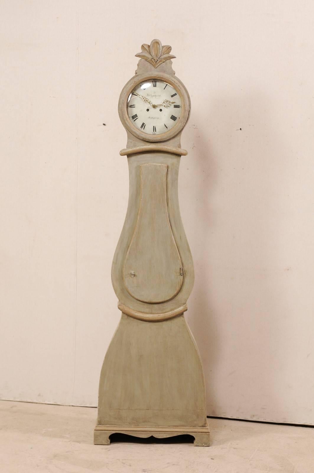 A Swedish 19th century painted wood floor clock. This antique floor clock from Sweden features a sweetly carved and raised fanned-leaf crest, atop the circular face with roman numerals and exquisitely adorn brass hands. The body has a teardrop