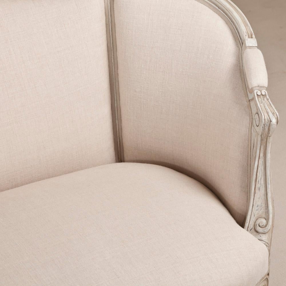 Un divano svedese dipinto di bianco/grigio con ispirazione rococò, dalle gambe cabriole, ai fiori e alle foglie intagliate, alla struttura a volute e alla forma curva del divano. Il divano è stato rivestito in un tessuto Nicole Fare color crema con