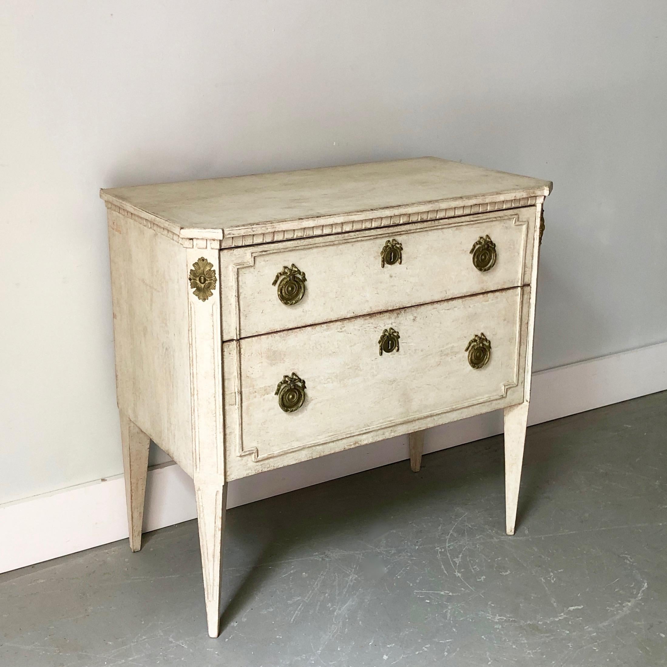 Charming Swedish two-drawer small chest, later paint in creamy white in Gustavian style with raised panels on the driver fronts, shaped top with dentil molding, canted corners, and long tapering legs. Bronze pulls and escutcheons.