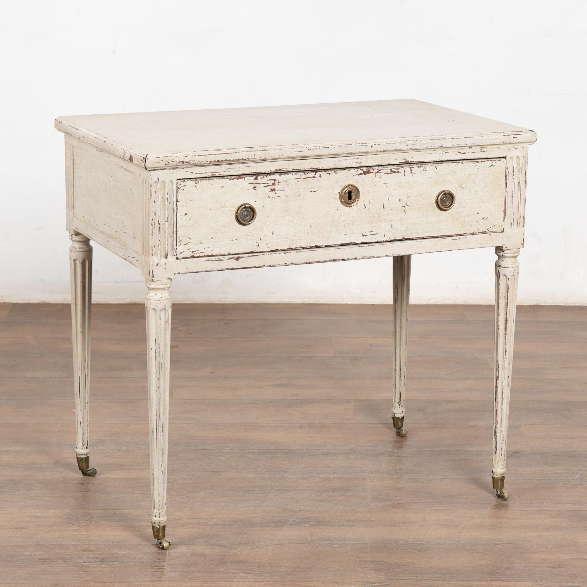 Antigua Mesa Auxiliar Sueca Gustaviana Pintada de Blanco con Cajón. Patas cónicas torneadas y estriadas con ruedas. También puede servir como mesilla de noche.
Restaurada, más tarde se aplicó profesionalmente un acabado de capas pintado en blanco