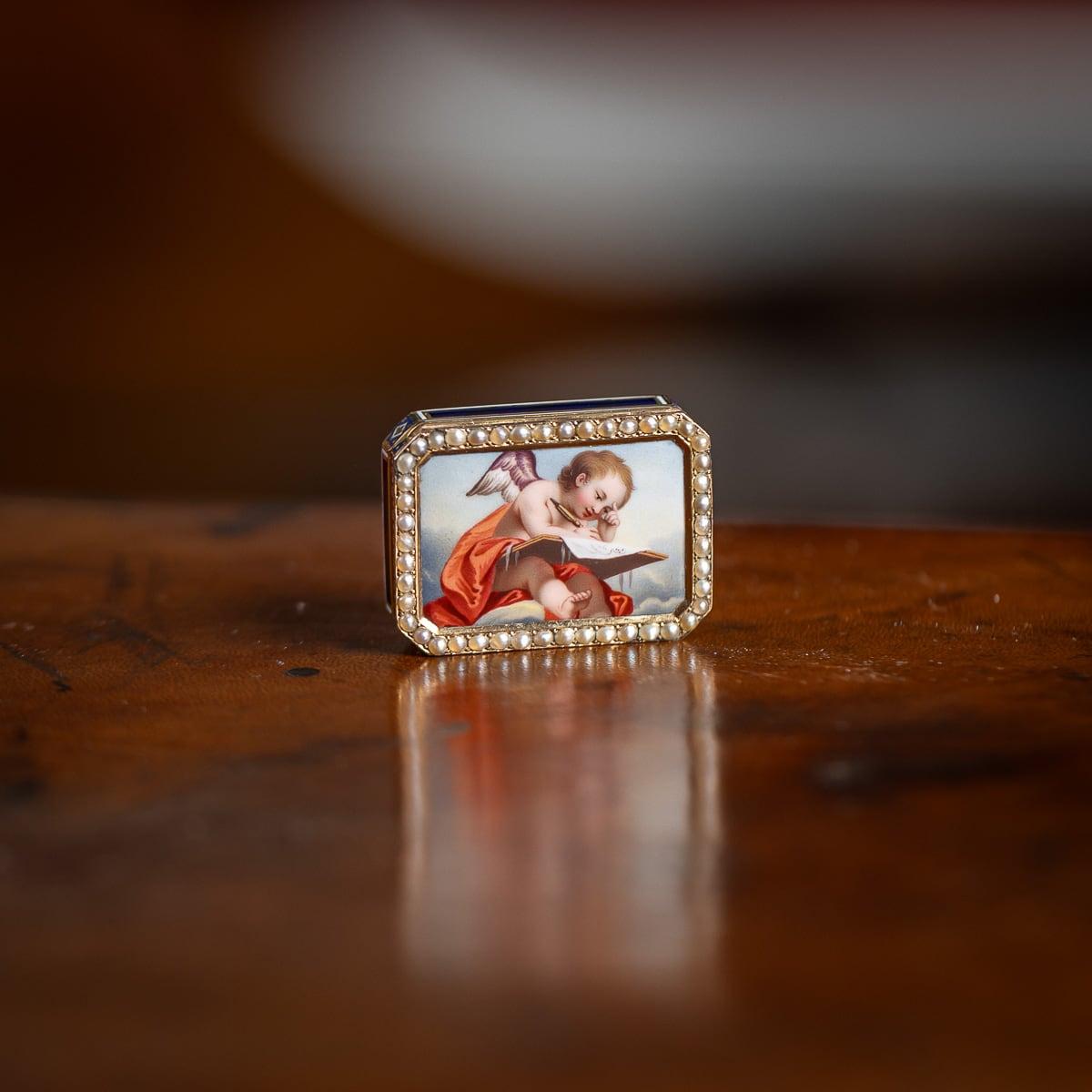 Vinaigrette ancienne du début du XIXe siècle en or suisse 18 carats, de forme rectangulaire avec des coins coupés, le couvercle représente un chérubin assis drapé de soie rouge, occupé à écrire, encadré par une délicate bordure de perles. Les côtés