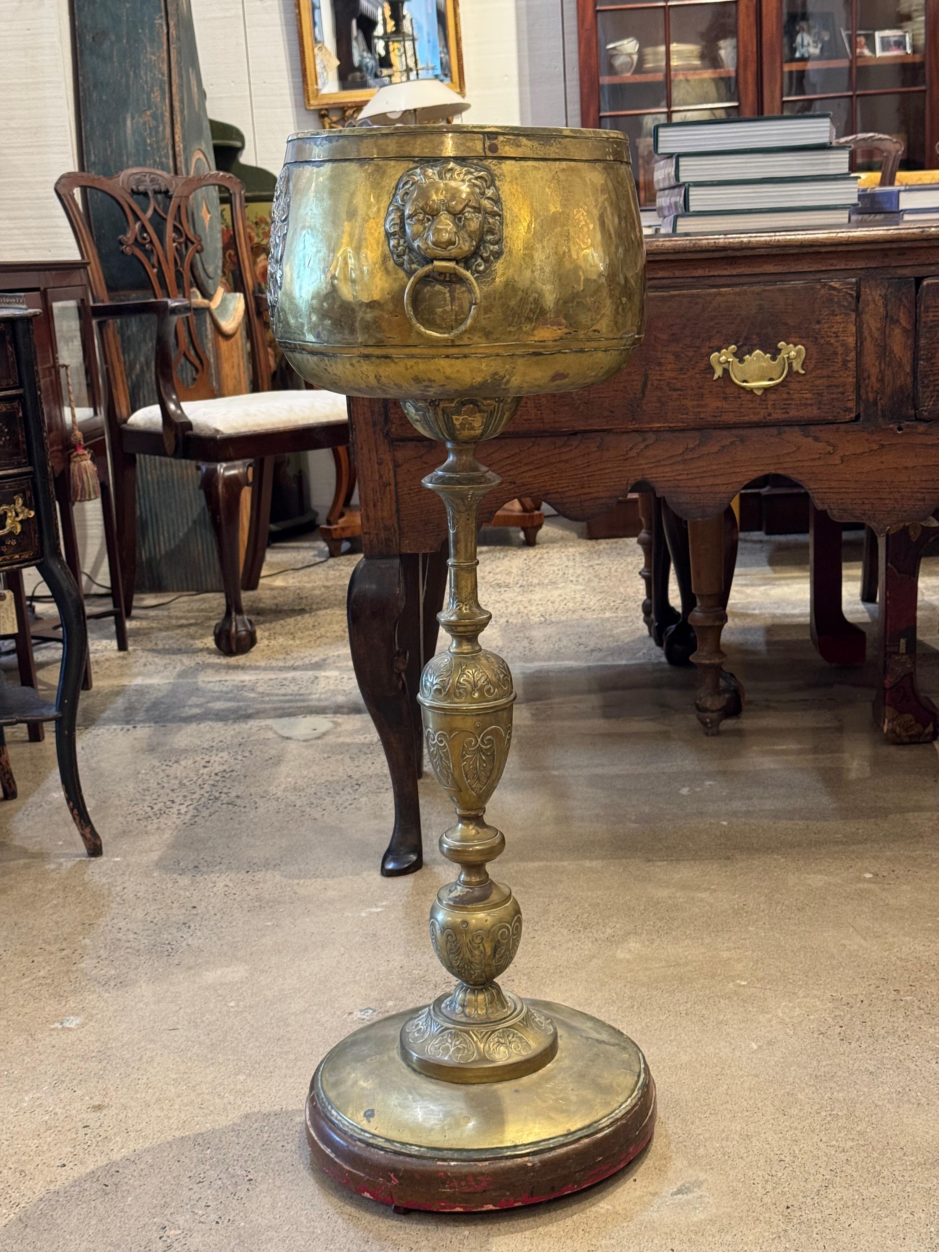 Vaso decorativo alto in ottone con base su piedistallo, dotato di manici ad anello con la classica testa di leone e dettagli incisi. Il fusto a colonna tornito e il piede svasato poggiano su una base circolare in legno con tracce di vernice rossa,