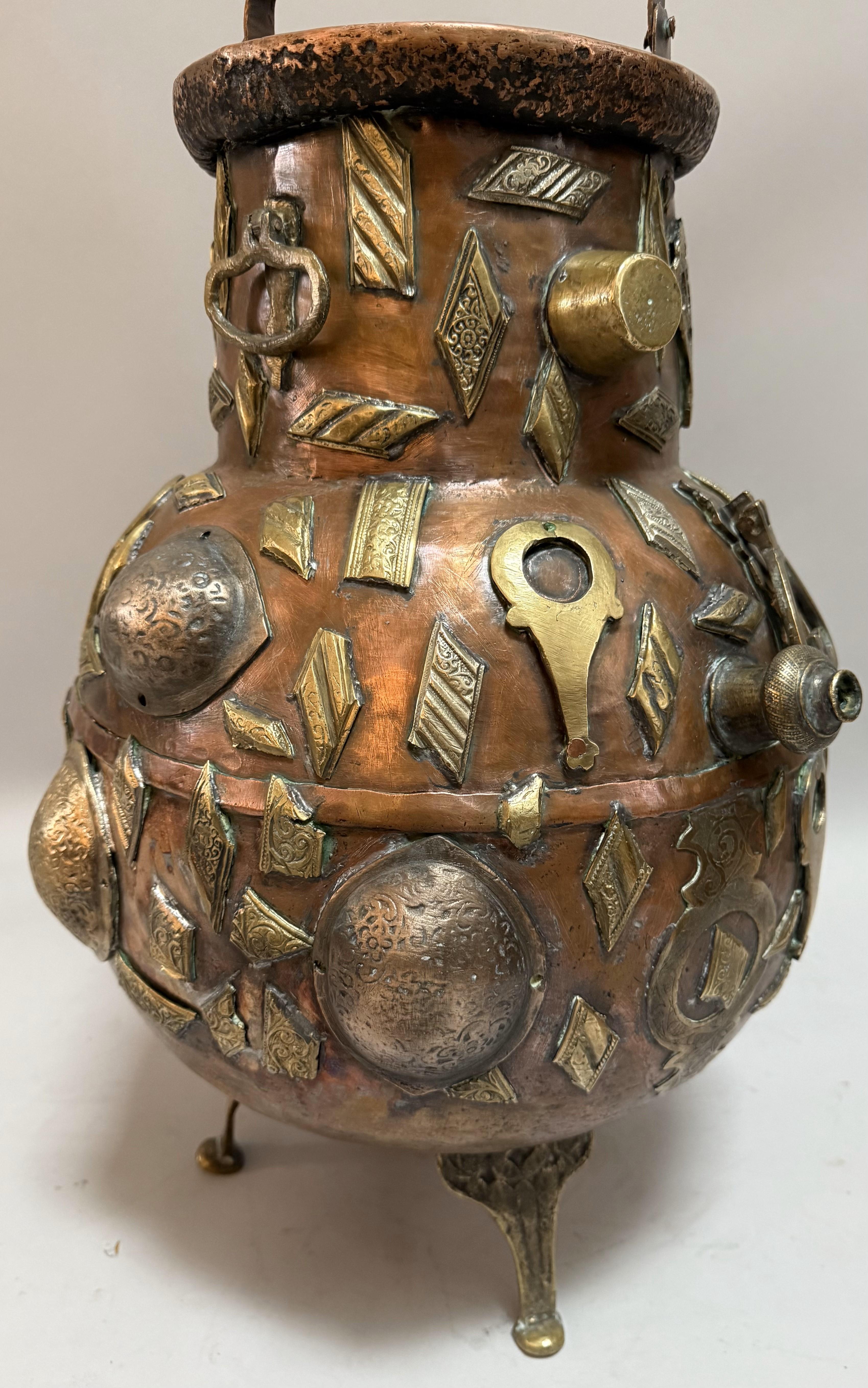 A richly detailed 19th-century Tibetan ritual vessel, hand-forged from copper and embellished with applied repousse and engraved brass medallions, geometric plaques, and symbolic mounts. Raised on three splayed feet, this bulbous, high-necked vessel