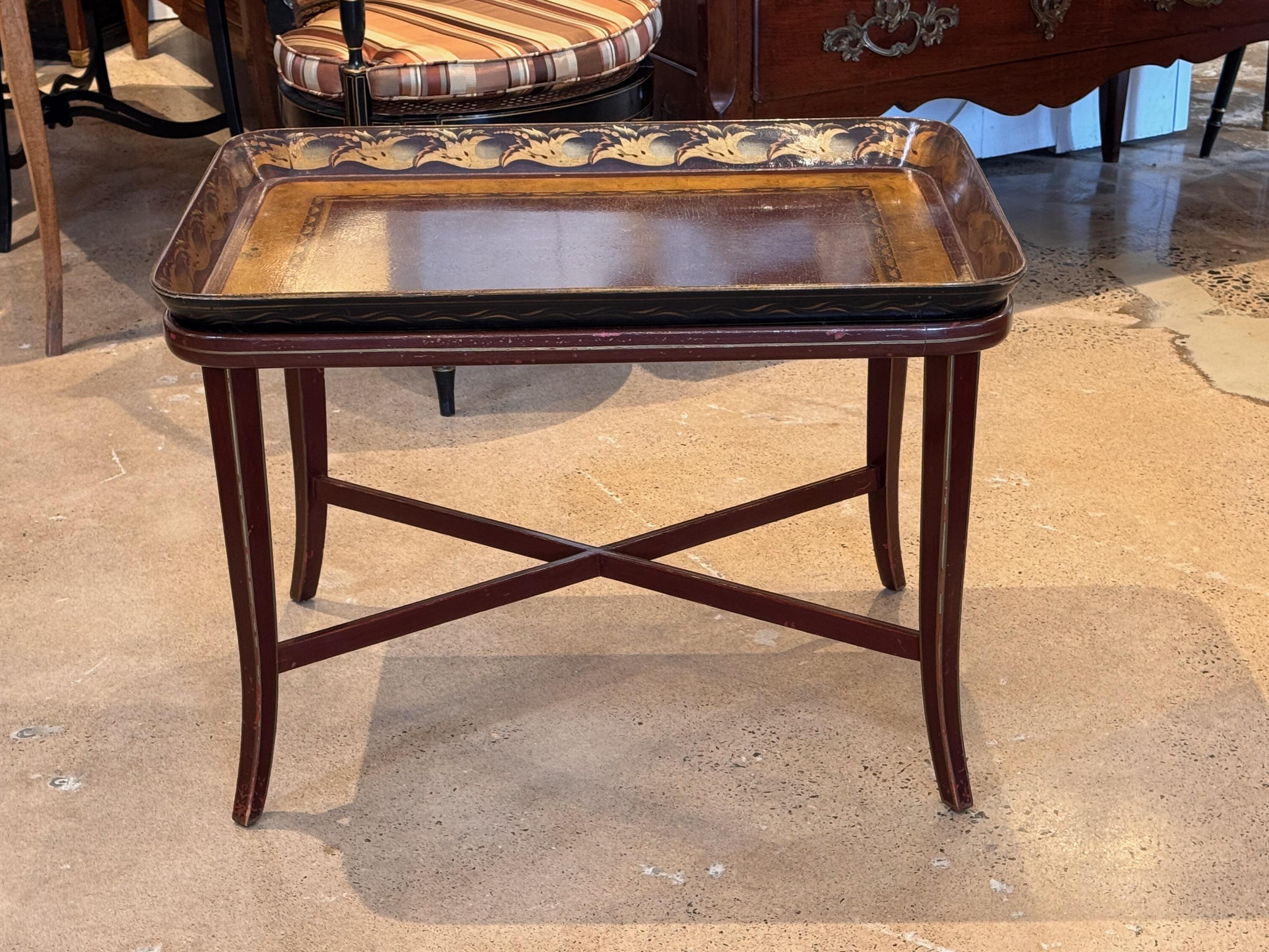 Cet élégant plateau antique sur pied est une pièce d'accent polyvalente et élégante, présentant un plateau magnifiquement peint à la main avec des détails dorés complexes sur le pourtour. La couleur de base bordeaux profond rehausse la richesse du