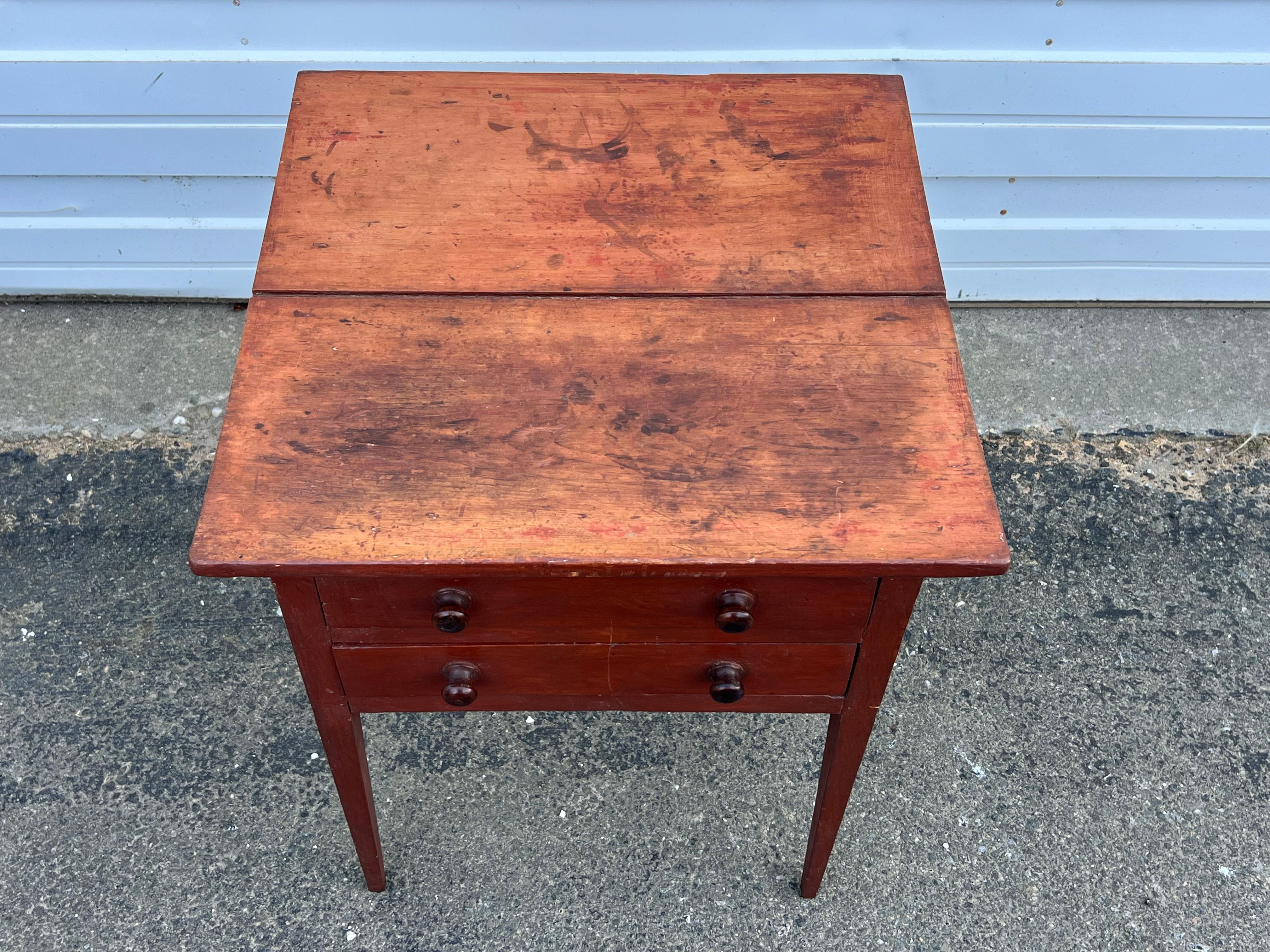 19th century two drawer side table in original red paint.  With two board, tongue and groove top, two drawers with turned wooden knobs, on tapered legs. 