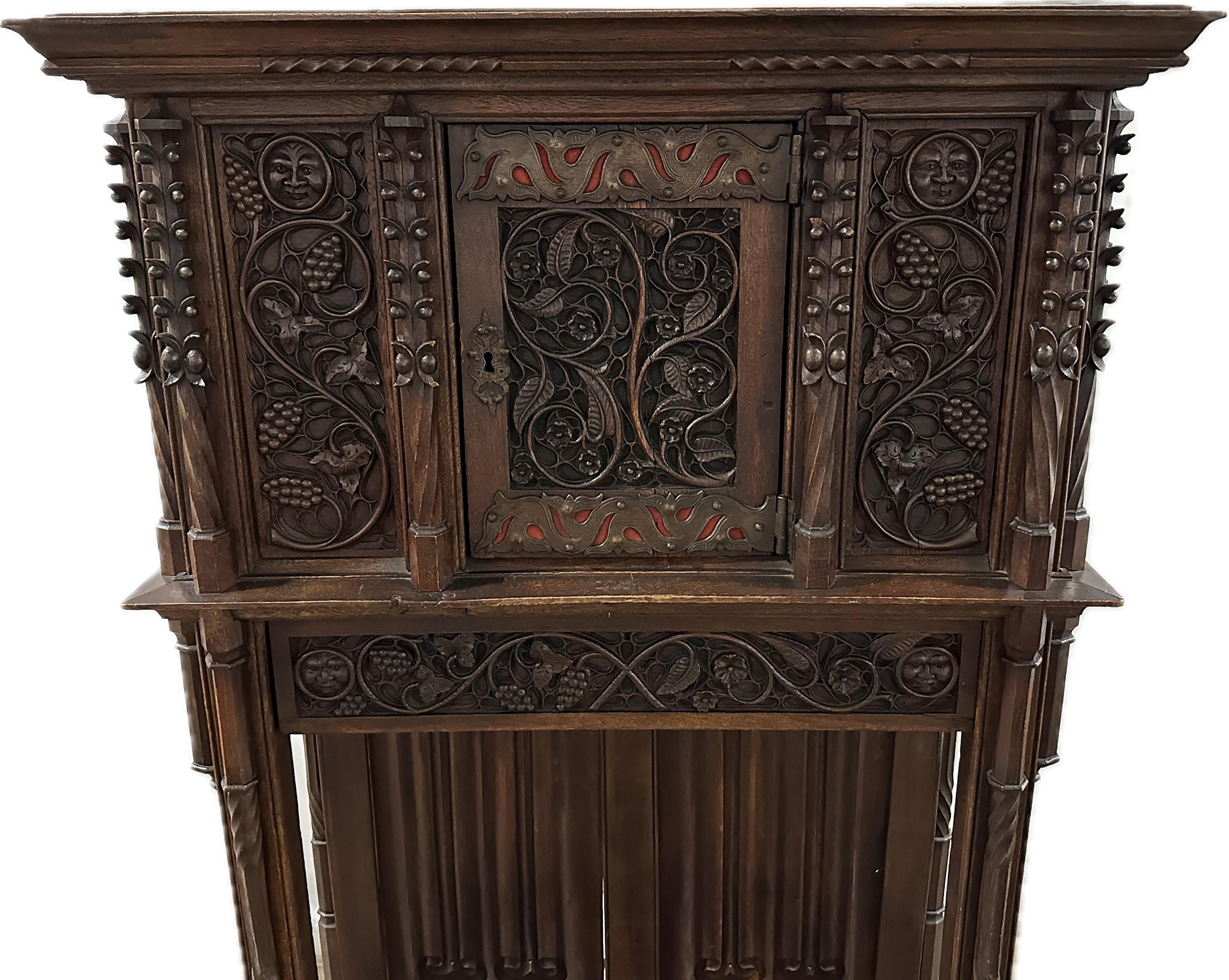 A handsome 19th Century two-piece gothic style oak cabinet. Beautifully carved and finished grapevines along the top and arms of the piece. A warm smiling face carving above the top of the vine carvings on either side of the front. Ventilated brass