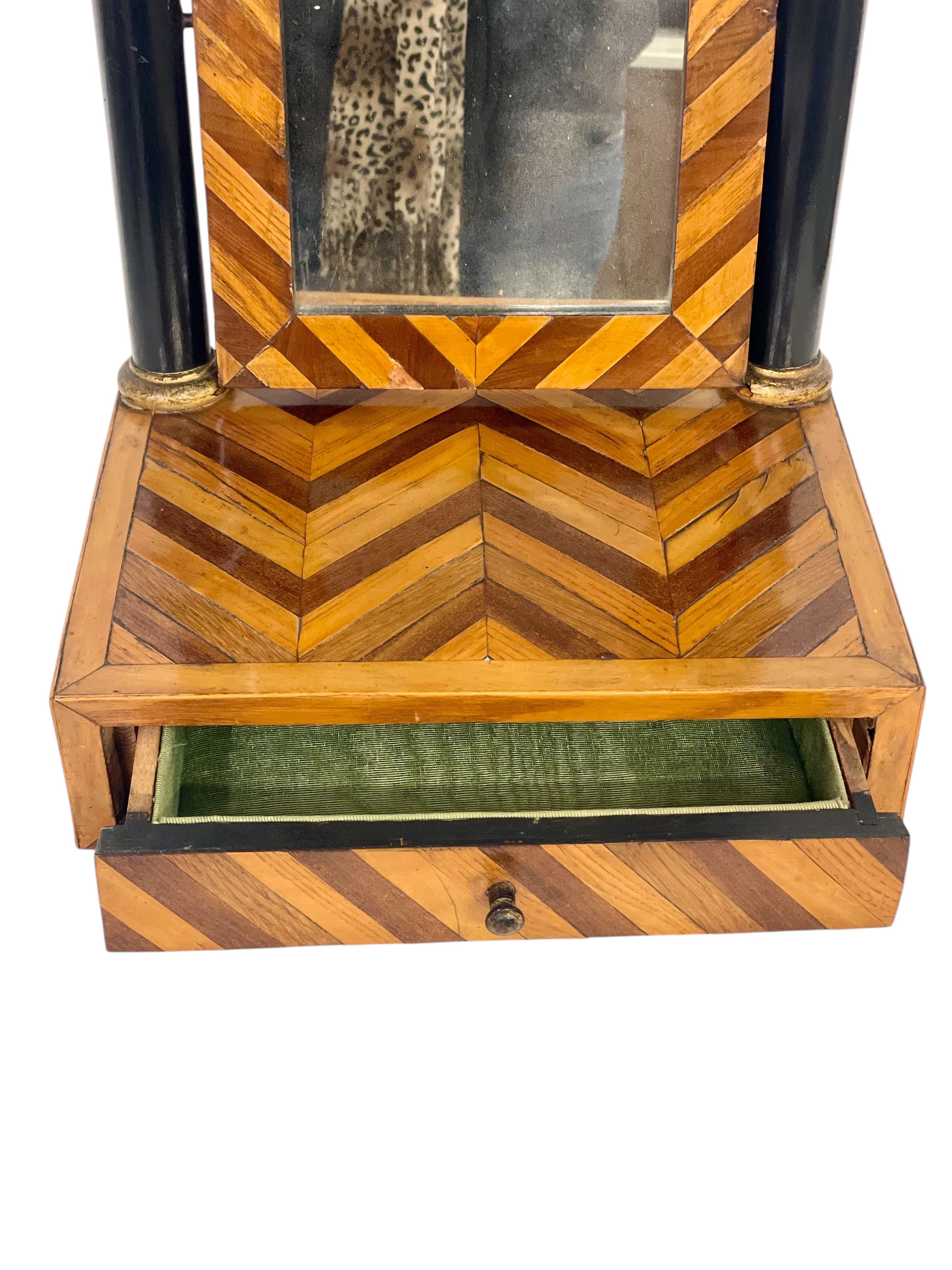 This charming early 19th-century French tabletop vanity mirror is a perfect blend of elegance and practicality. The chevron-patterned veneer frame highlights the craftsmanship of the period, while the ebonized columns with gilded accents add a touch