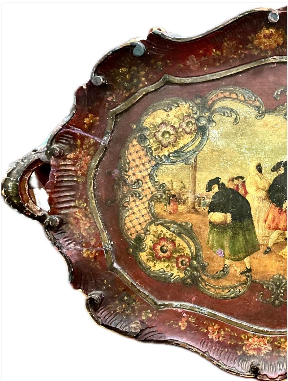 Bandeja de madera lacada veneciana del siglo XIX. Bandeja con decoración floral y una escena rural con figuras que representan la vida veneciana, todo sobre fondo burdeos. La bandeja presenta un bello festoneado ornamental en los bordes y dos asas,